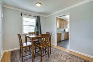 A dining area with a wooden table and chairs, a window with curtains, and a view into the kitchen.