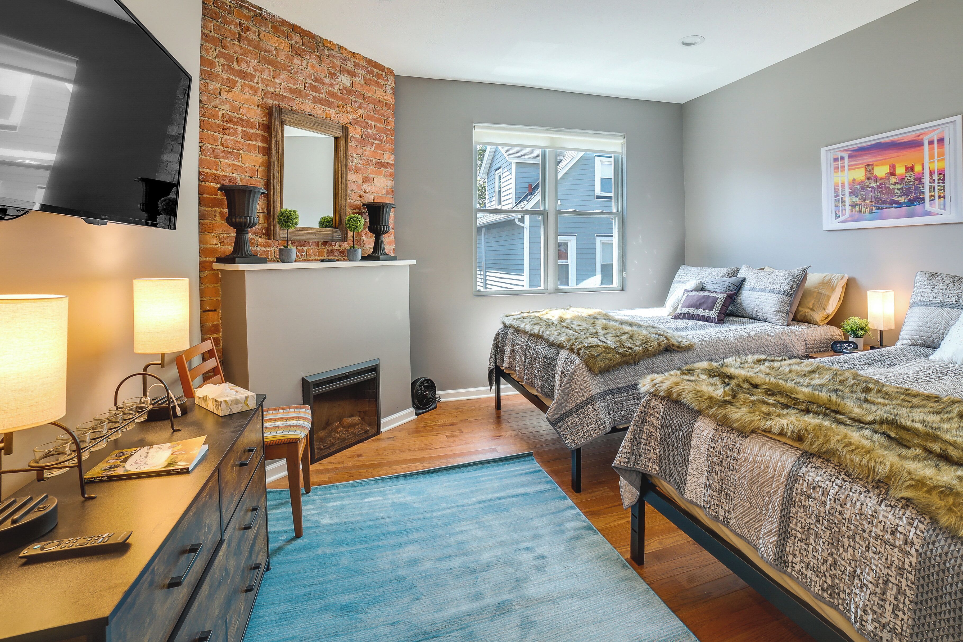 A bedroom with two beds, a brick accent wall, a fireplace, and a large window with a city view.
