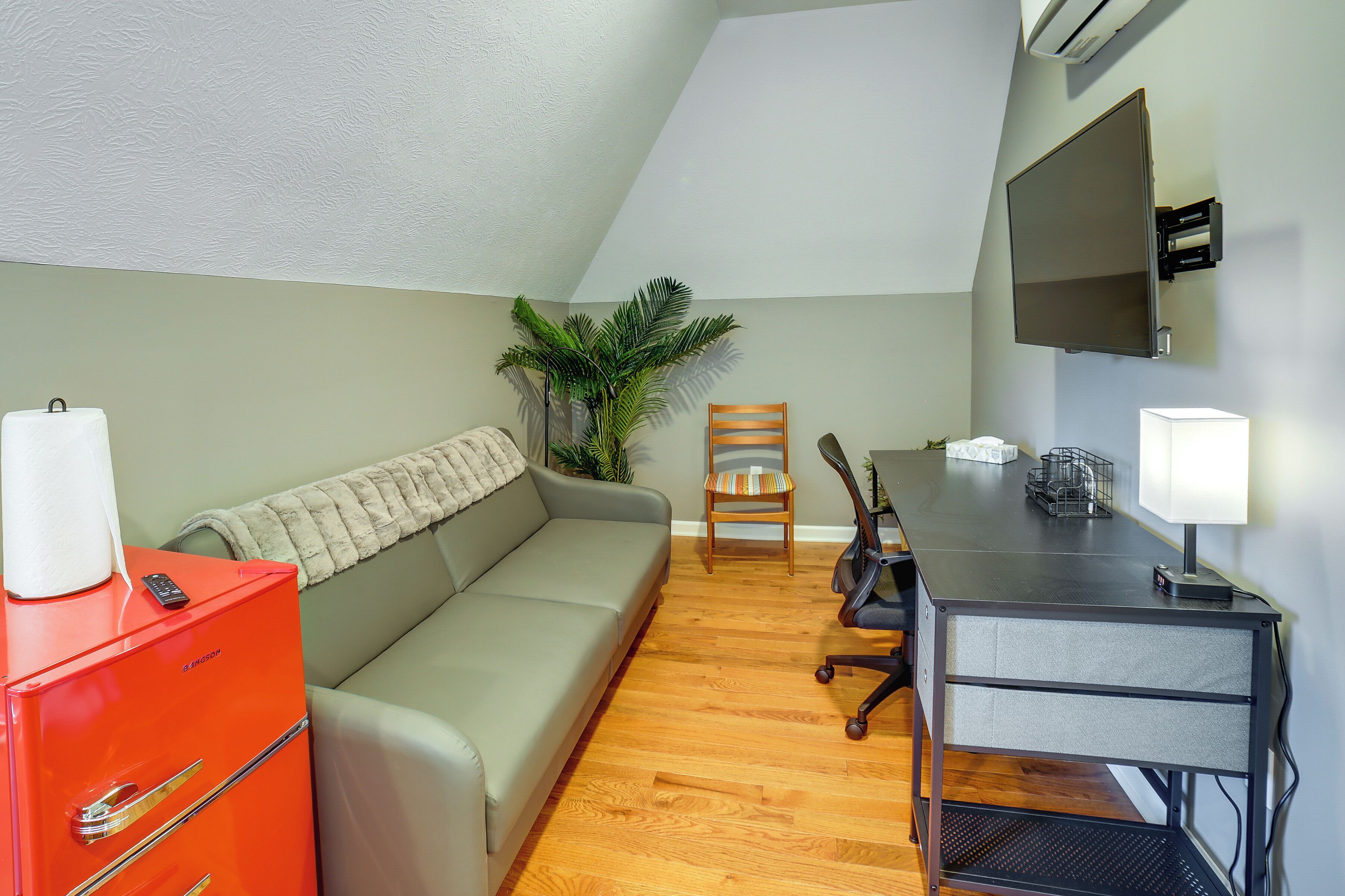 Un espacio habitable compacto con sofá, una mesita, una silla, un armario rojo, una planta en maceta y un televisor montado.