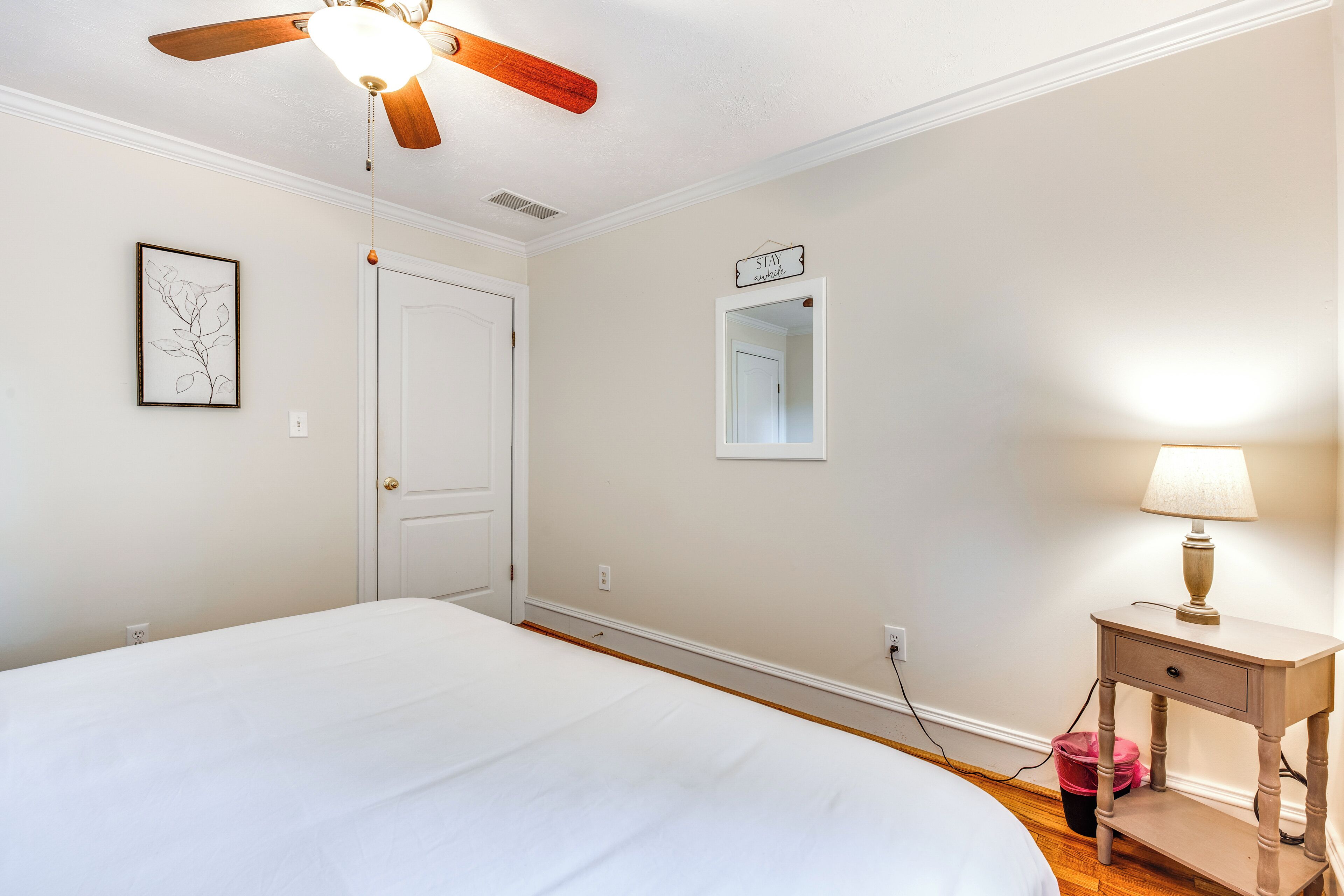 A bedroom with a bed, a ceiling fan, a nightstand with a lamp, a door, and a framed picture on the wall.