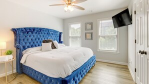 A bedroom with a tufted blue headboard, a flat-screen TV mounted on the wall, and a ceiling fan. - Ideal ATL Base 2 Mi to Mercedes-benz Stadium, Dtwn (Atlanta)