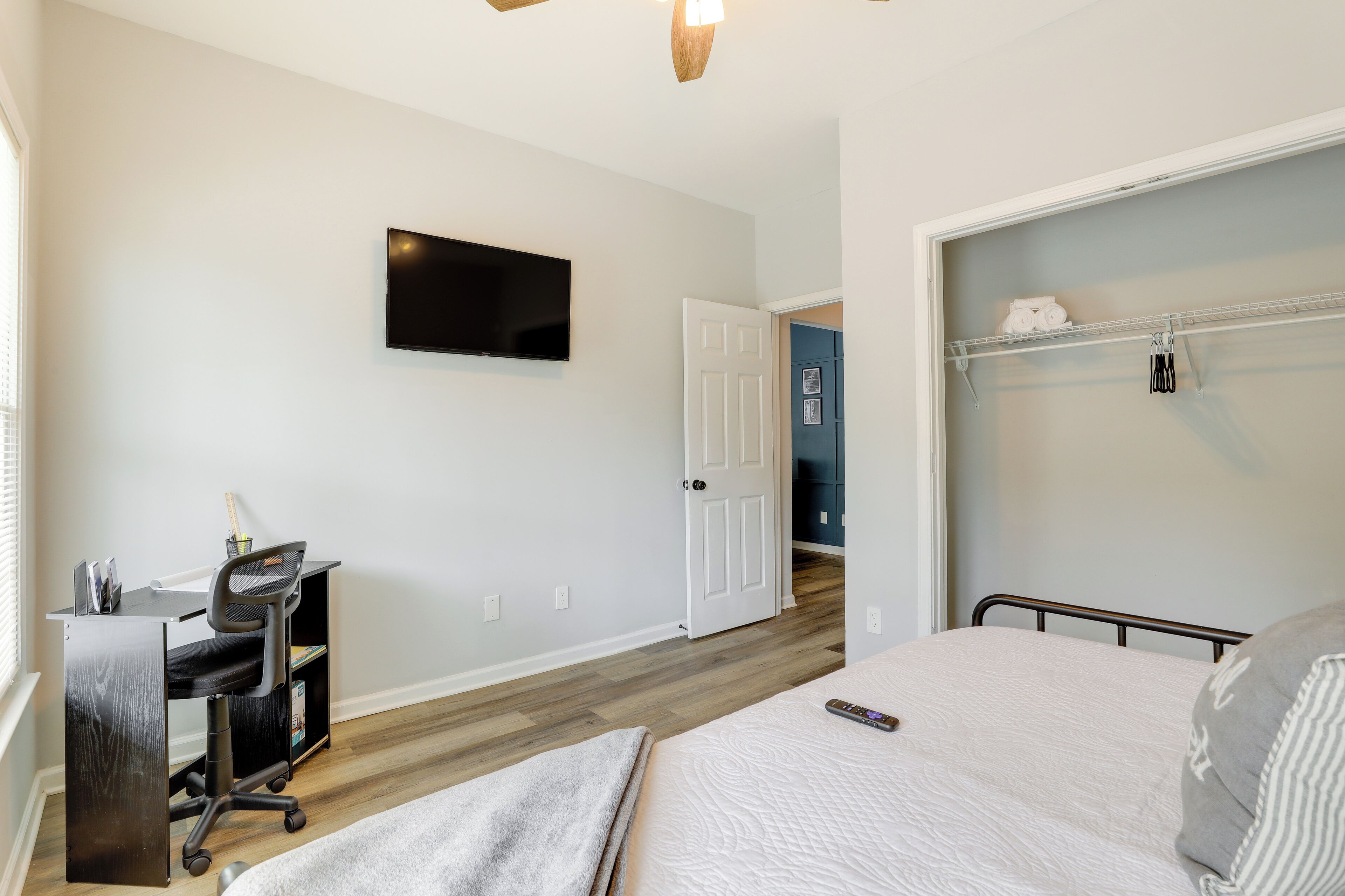 A bedroom with a bed, a desk with a chair, a television on the wall, and a door leading to another room.