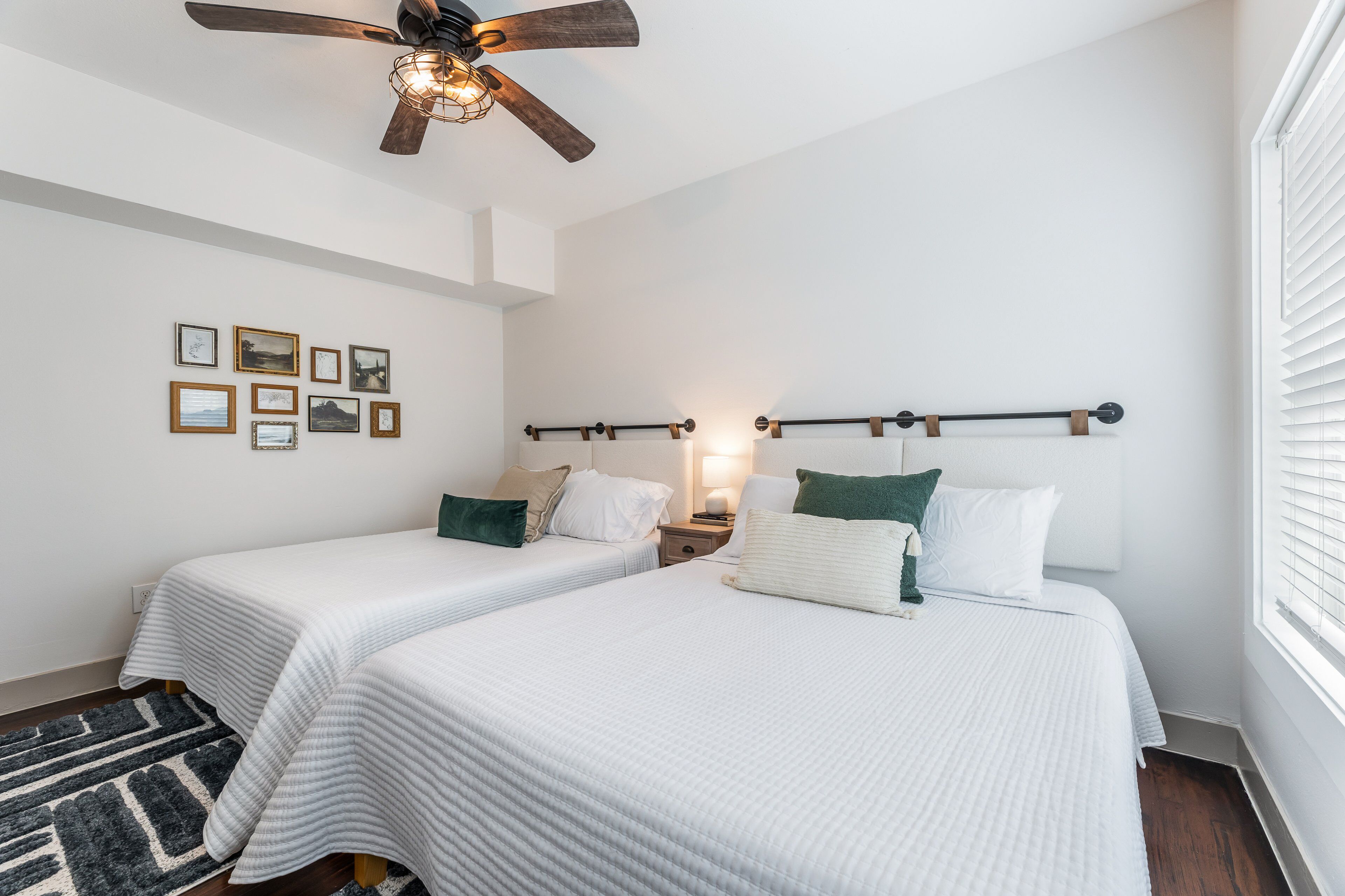 A bedroom with two beds, a ceiling fan, wall-mounted lights, a rug, and framed pictures.