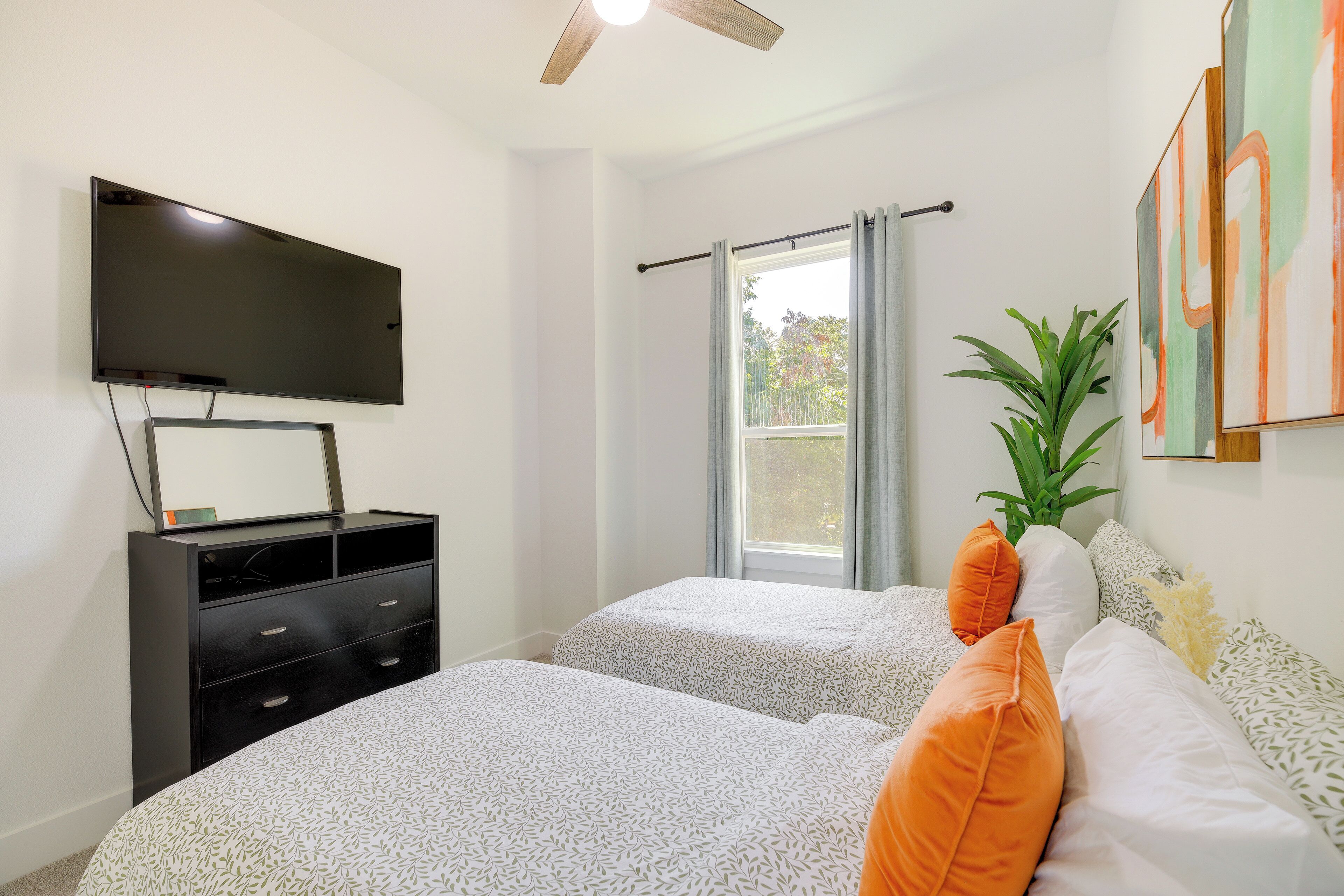 Une chambre avec deux lits, une télévision, un ventilateur de plafond, une fenêtre avec des rideaux et une plante.