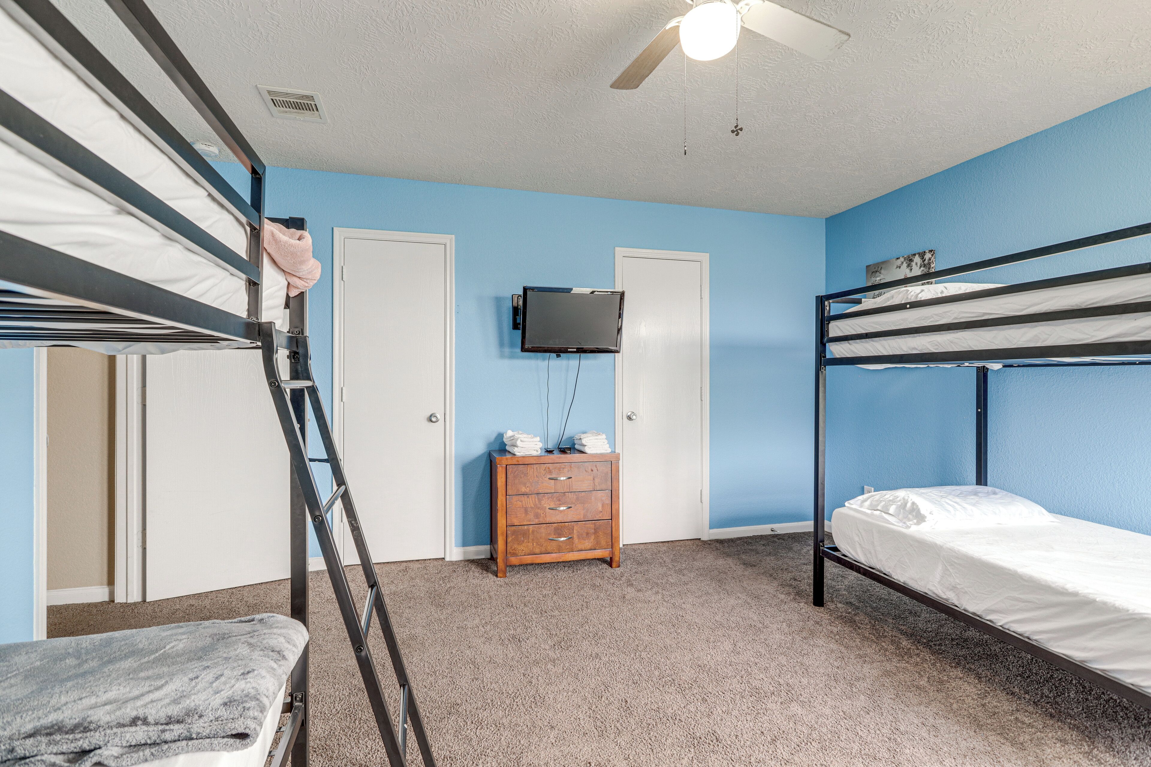 A bunk bed room with a ceiling fan, a TV mounted on the wall, and a wooden dresser.
