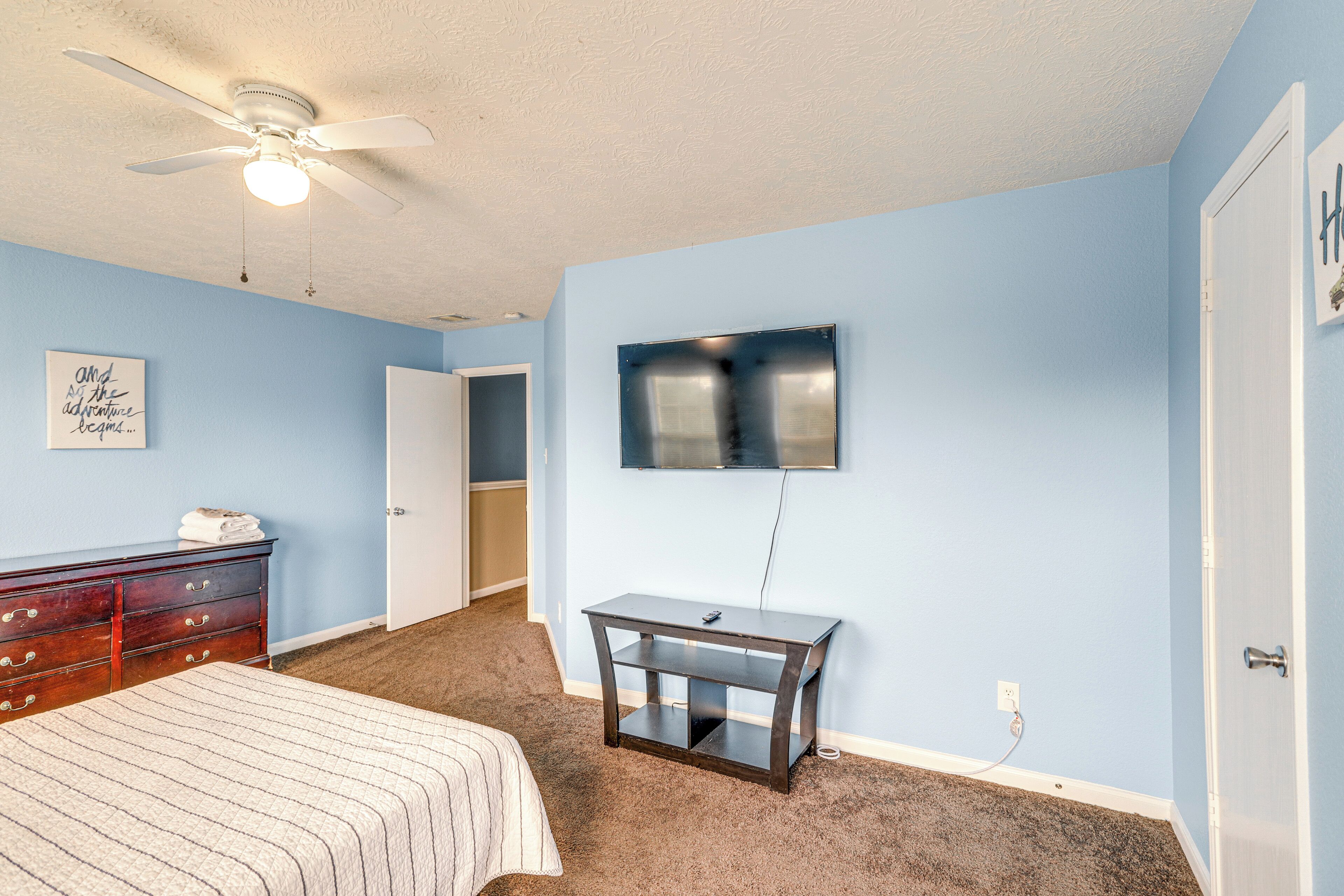 A bedroom with a bed, a dresser, a desk, a ceiling fan, and a TV mounted on the wall.