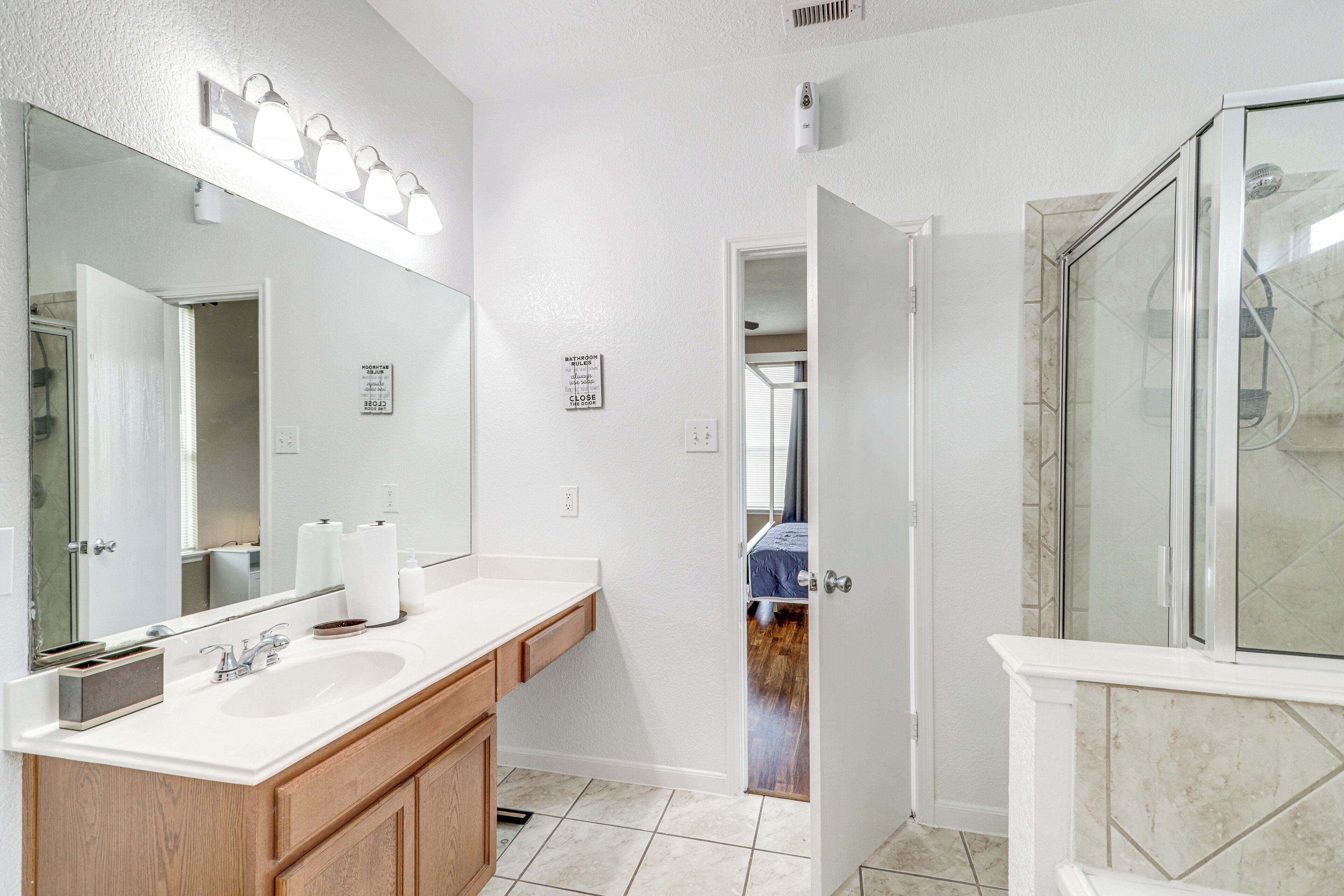A bathroom with a large mirror, two sinks, a shower, and a door leading to another room.