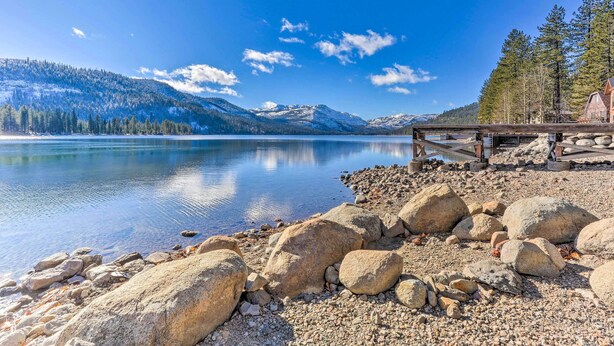A lakeside cabin with a wooden deck overlooking a calm lake, surrounded by mountains and trees.