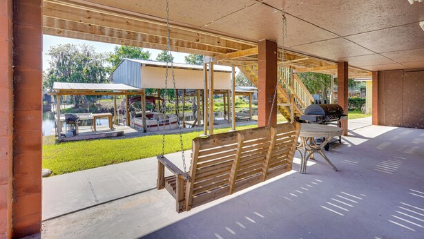 A covered patio area with a swing set, a barbecue grill, and a view of a park with a boat and a building.