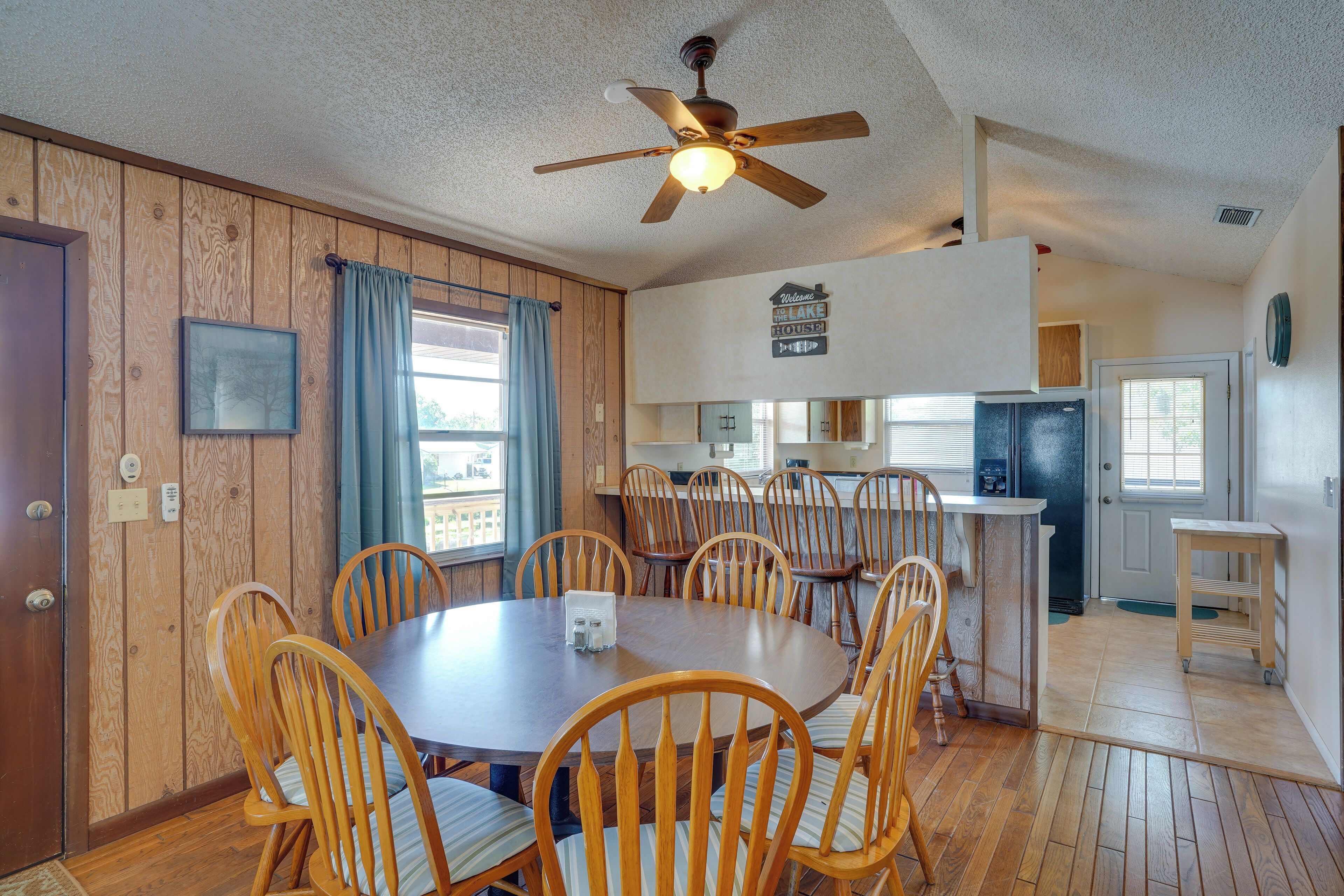 A dining area with a round table and wooden chairs, a ceiling fan, and a kitchen in the background.