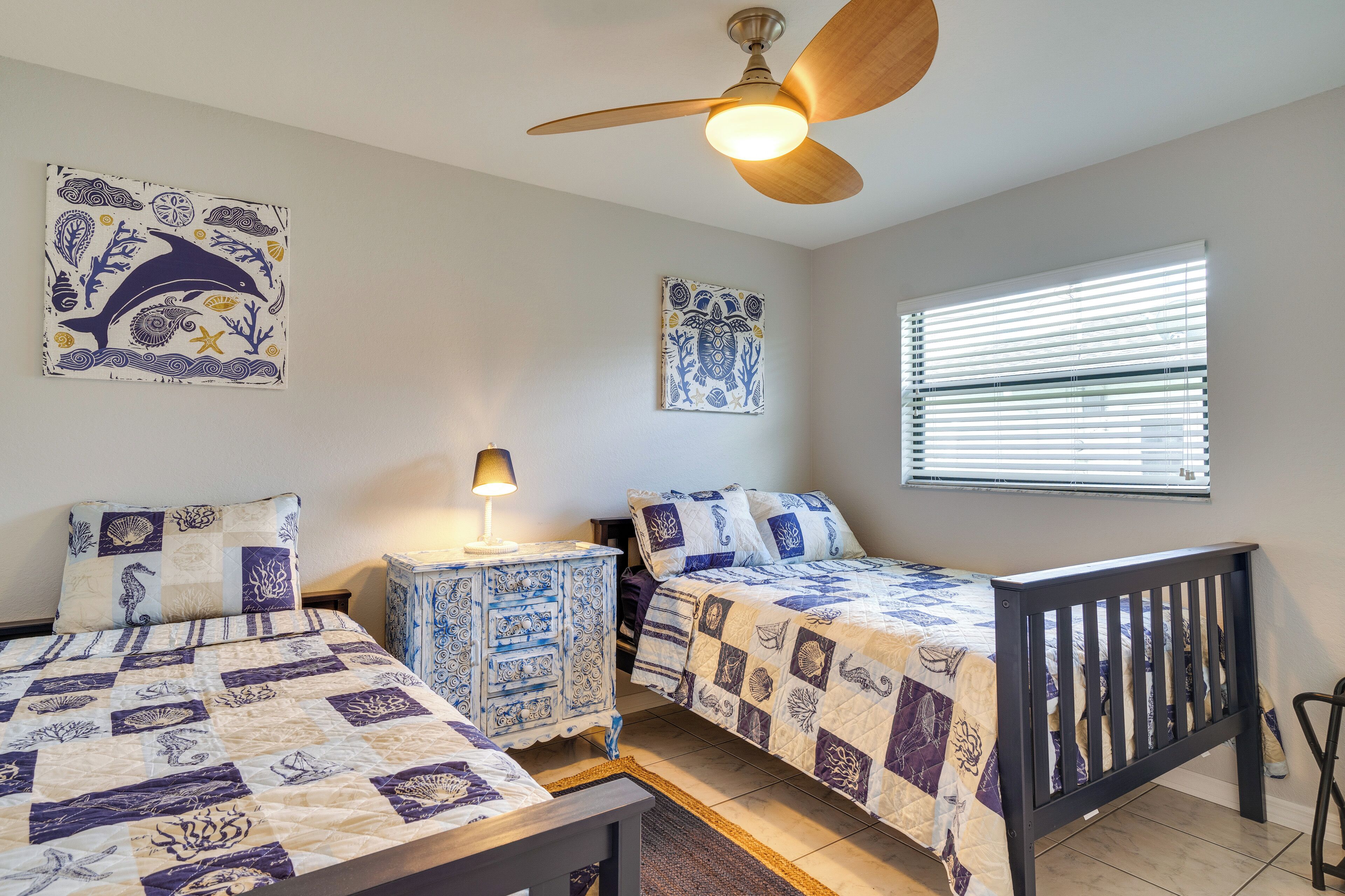 A bedroom with two beds, a ceiling fan, a window with blinds, and decorative wall art.