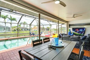 A poolside dining area with a table, chairs, and a TV screen. - Lanai & Pool: Waterfront Cape Coral Home! (Cape Coral)