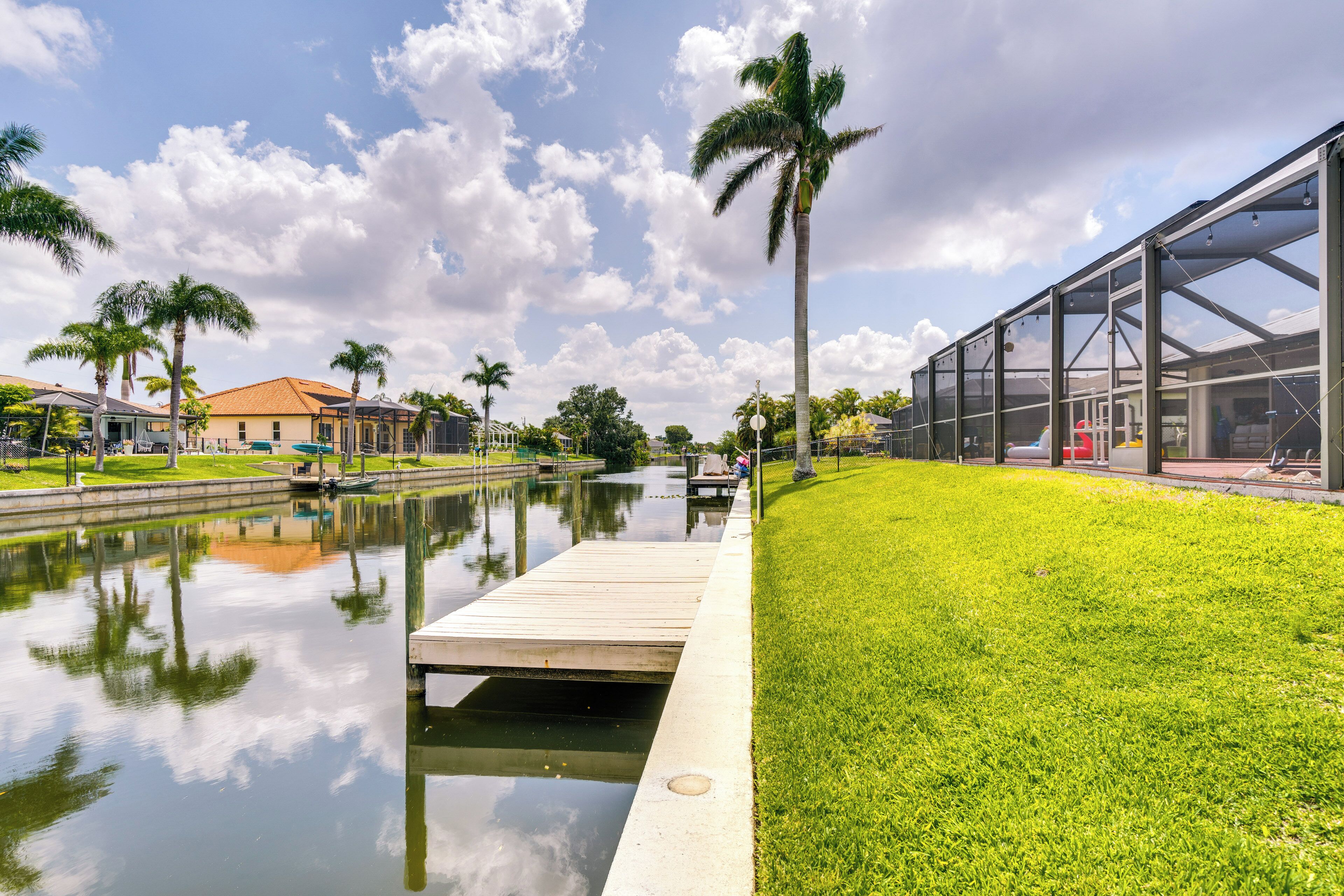 A waterfront park with a wooden dock, palm trees, and a modern building with large windows.