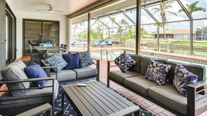 Un amplio espacio exterior con piscina, ventilador de techo y grandes ventanales que ofrecen vistas a palmeras y una zona junto a la piscina.