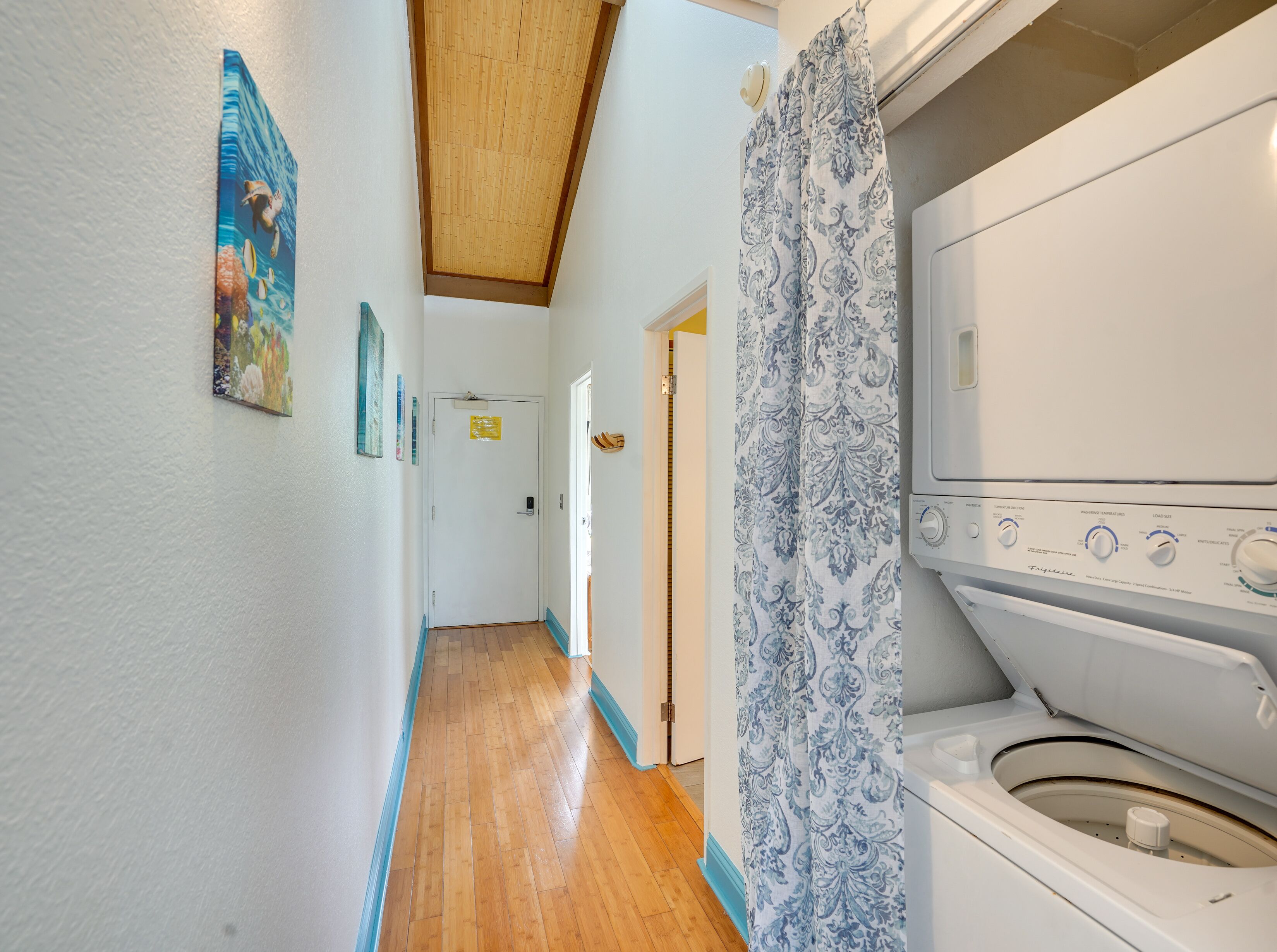 A laundry room with a front-loading washer and dryer, wooden flooring, and a hallway with a door leading to another room.