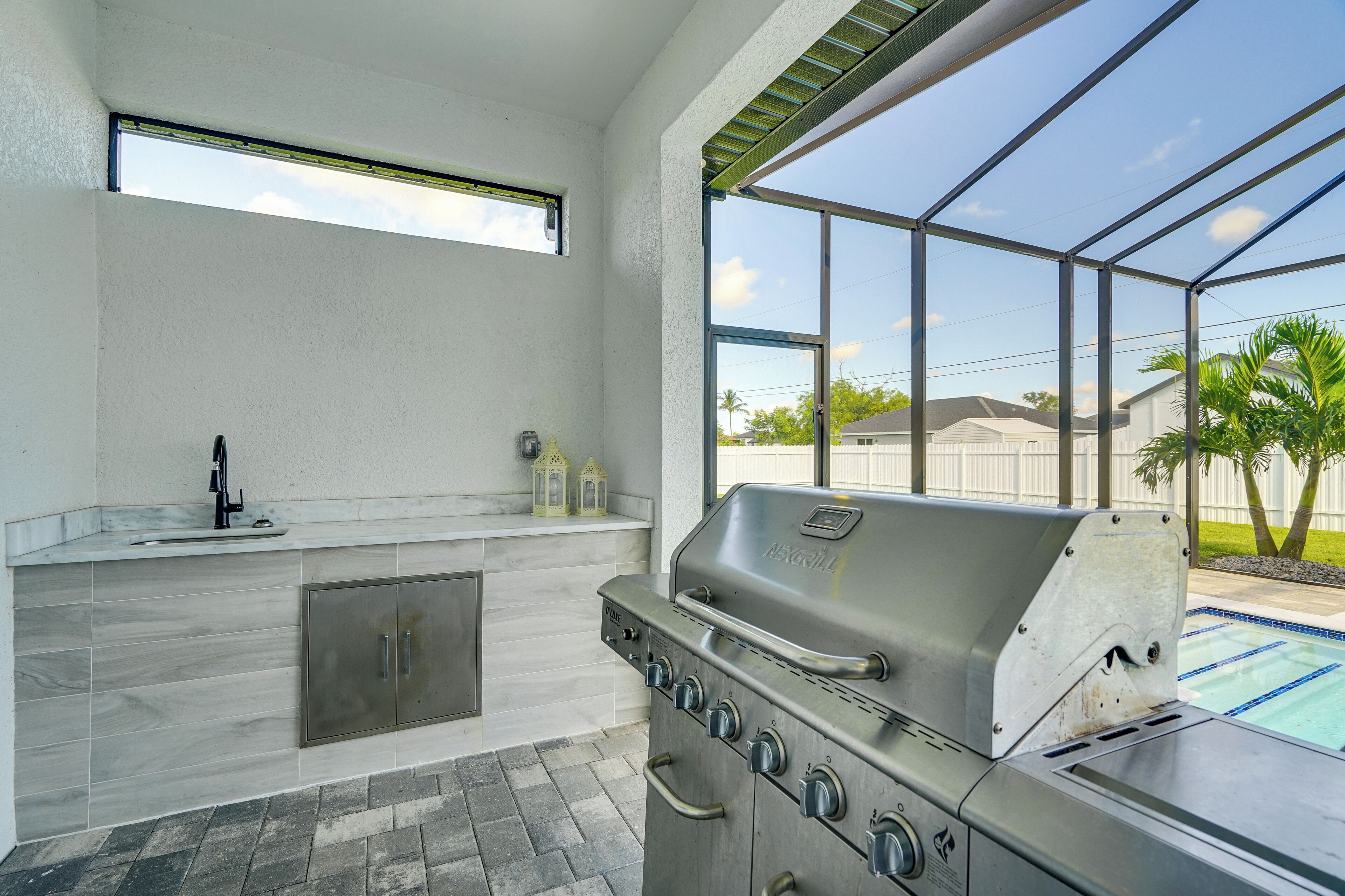 Una cocina exterior moderna con una parrilla empotrada, un lavamanos y una ventana con vistas a una piscina.