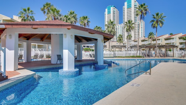 A pool area with a covered seating area, palm trees, and high-rise buildings in the background. - South Padre Island Condo: Walk to the Beach! (South Padre Island)