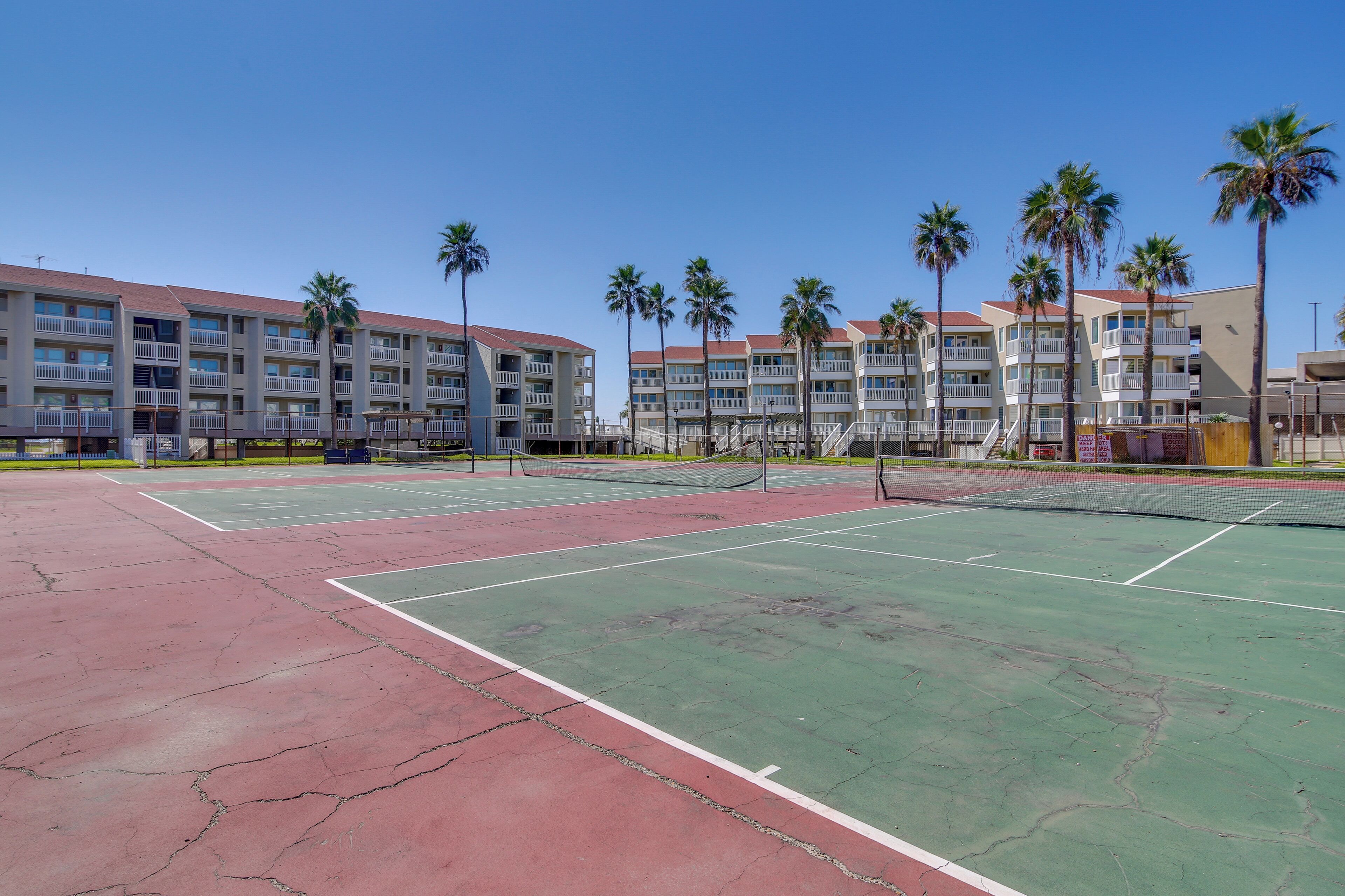 A tennis court with a net, surrounded by palm trees and a multi-story building with balconies.
