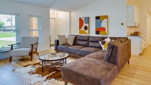 A modern living room with a brown leather sofa, a patterned armchair, and a glass coffee table. There are abstract paintings on the wall and a kitchen area in the background. - Heated Pool: Pet-friendly Home in Cape Coral (Cape Coral)