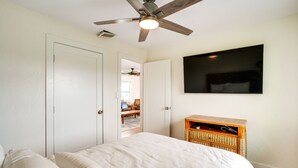 Une chambre à coucher avec un lit, un ventilateur de plafond, un téléviseur fixé au mur et une table d’appoint en osier.