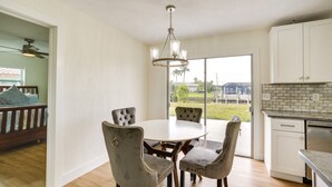Un coin repas avec une table ronde et quatre chaises, une cuisine avec des meubles blancs et un lave-vaisselle, et une vue sur un espace verdoyant accessible par une porte-fenêtre coulissante.