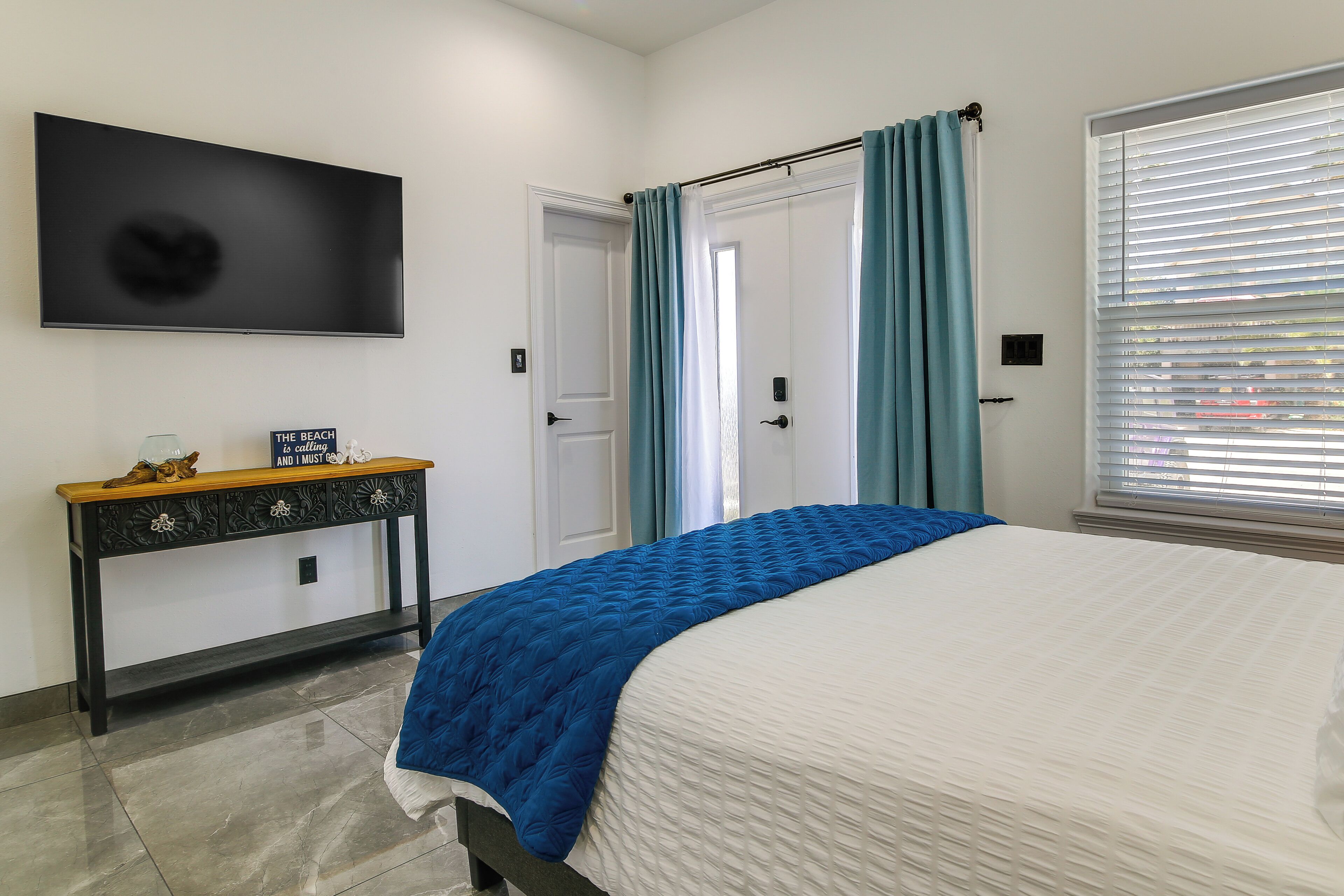 Une chambre à coucher avec un lit, un téléviseur fixé au mur, une console sur laquelle est posé un panneau, et une fenêtre avec des stores.