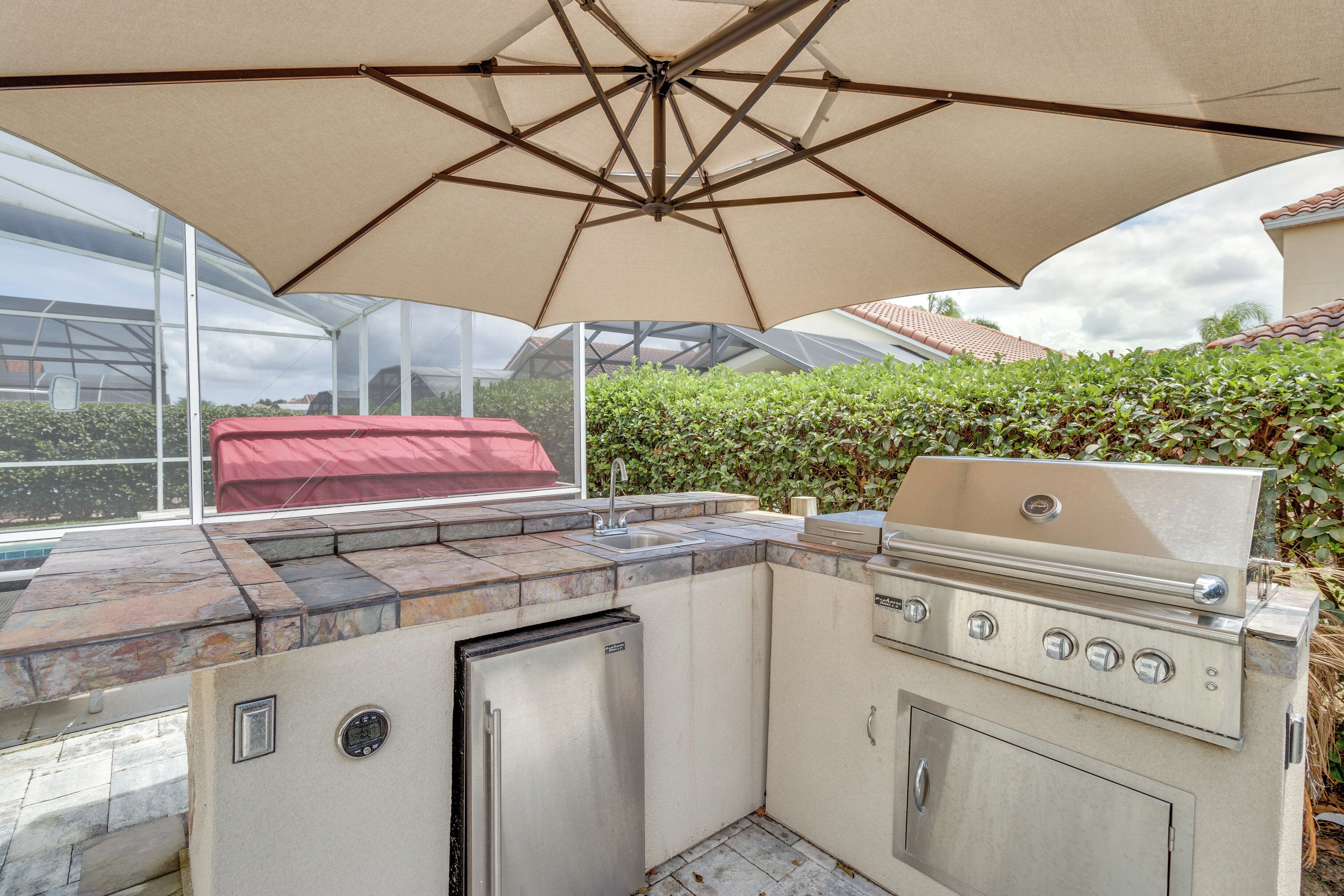 Une cuisine extérieure équipée d’un barbecue, d’un évier et d’un réfrigérateur, située sous un grand parasol.