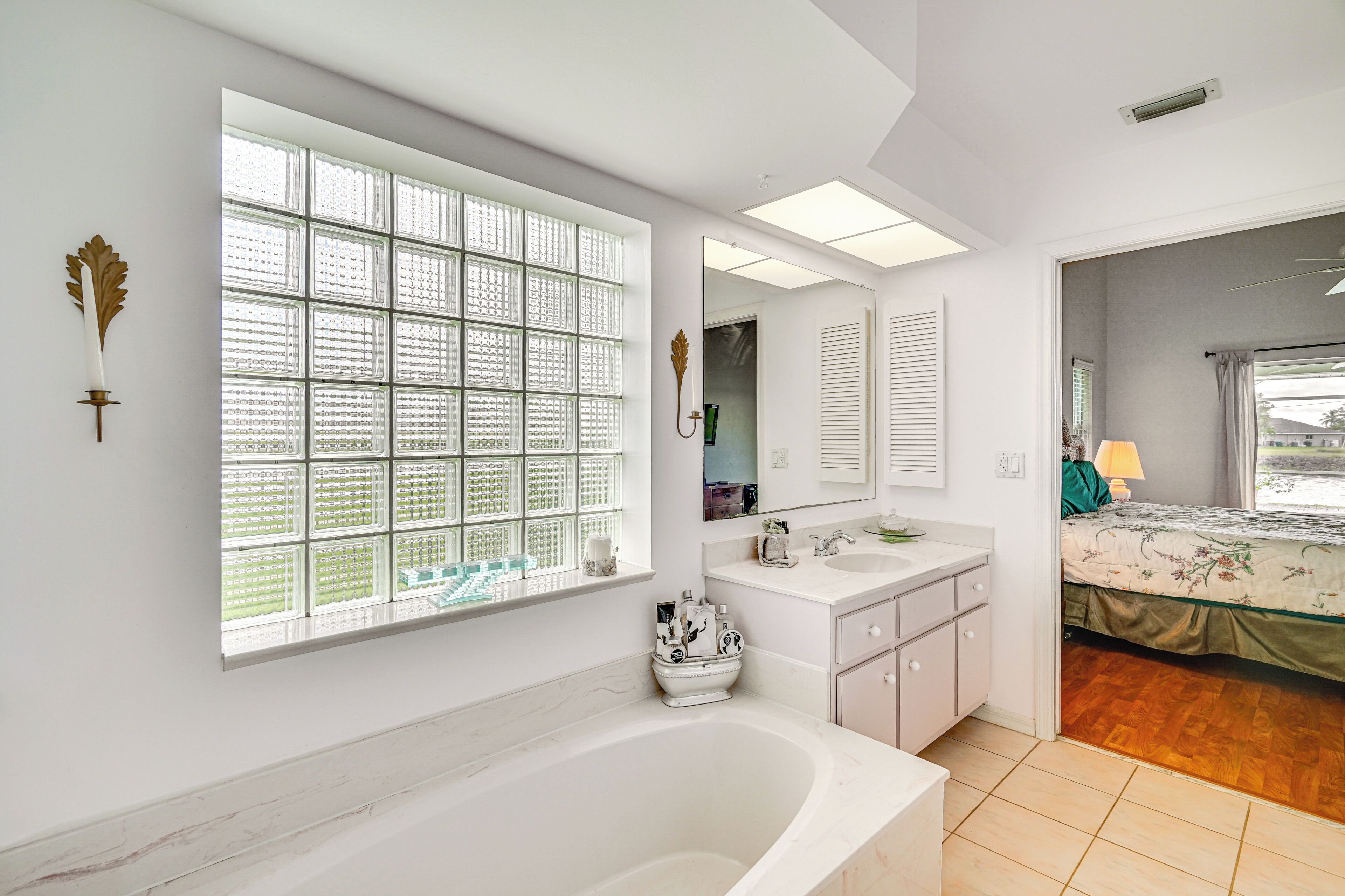A modern bathroom with a large window, a bathtub, a vanity with a mirror, and a bed in the adjacent room.
