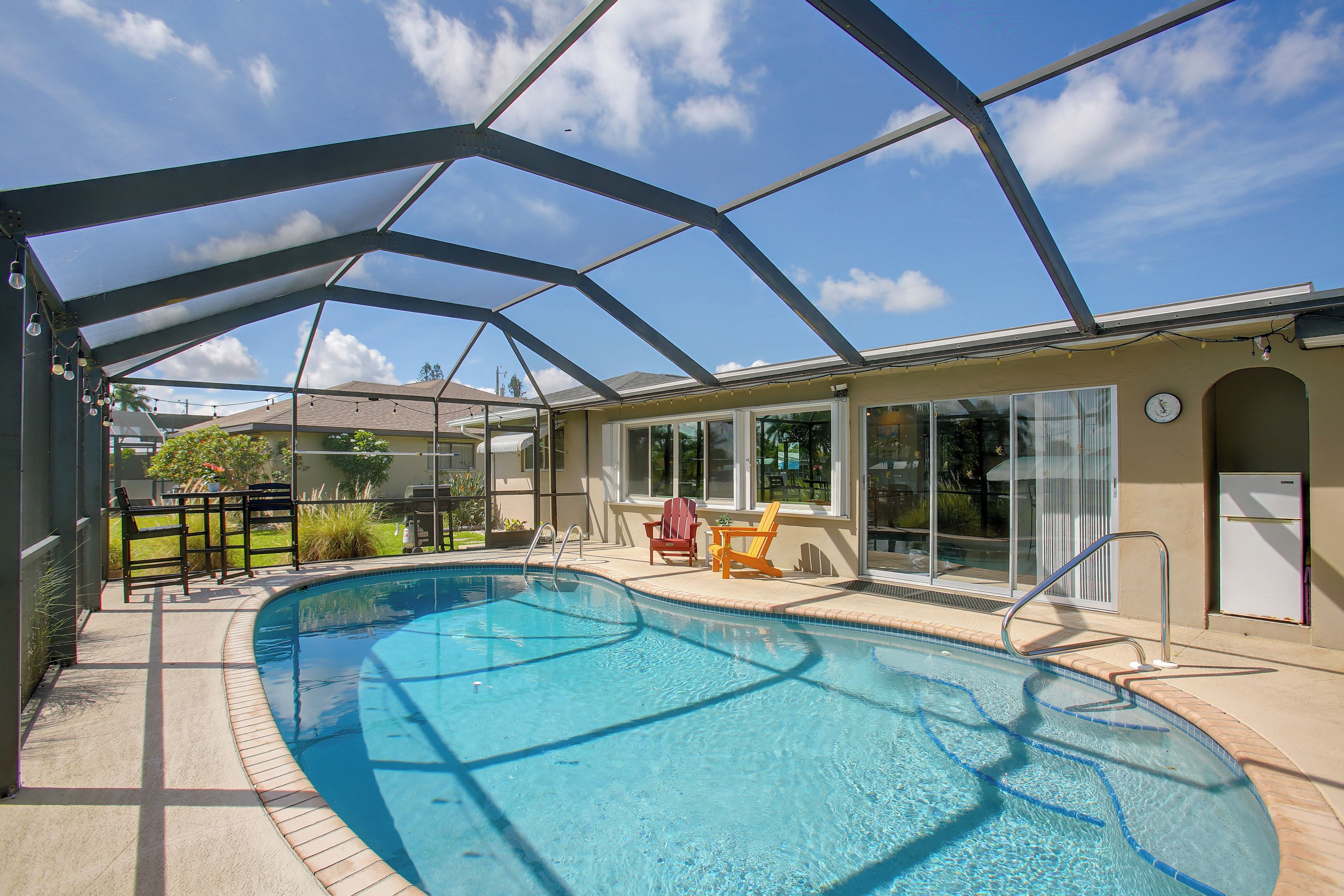 A covered swimming pool with a glass enclosure, adjacent to a building with a sliding glass door, and a patio area with outdoor furniture.