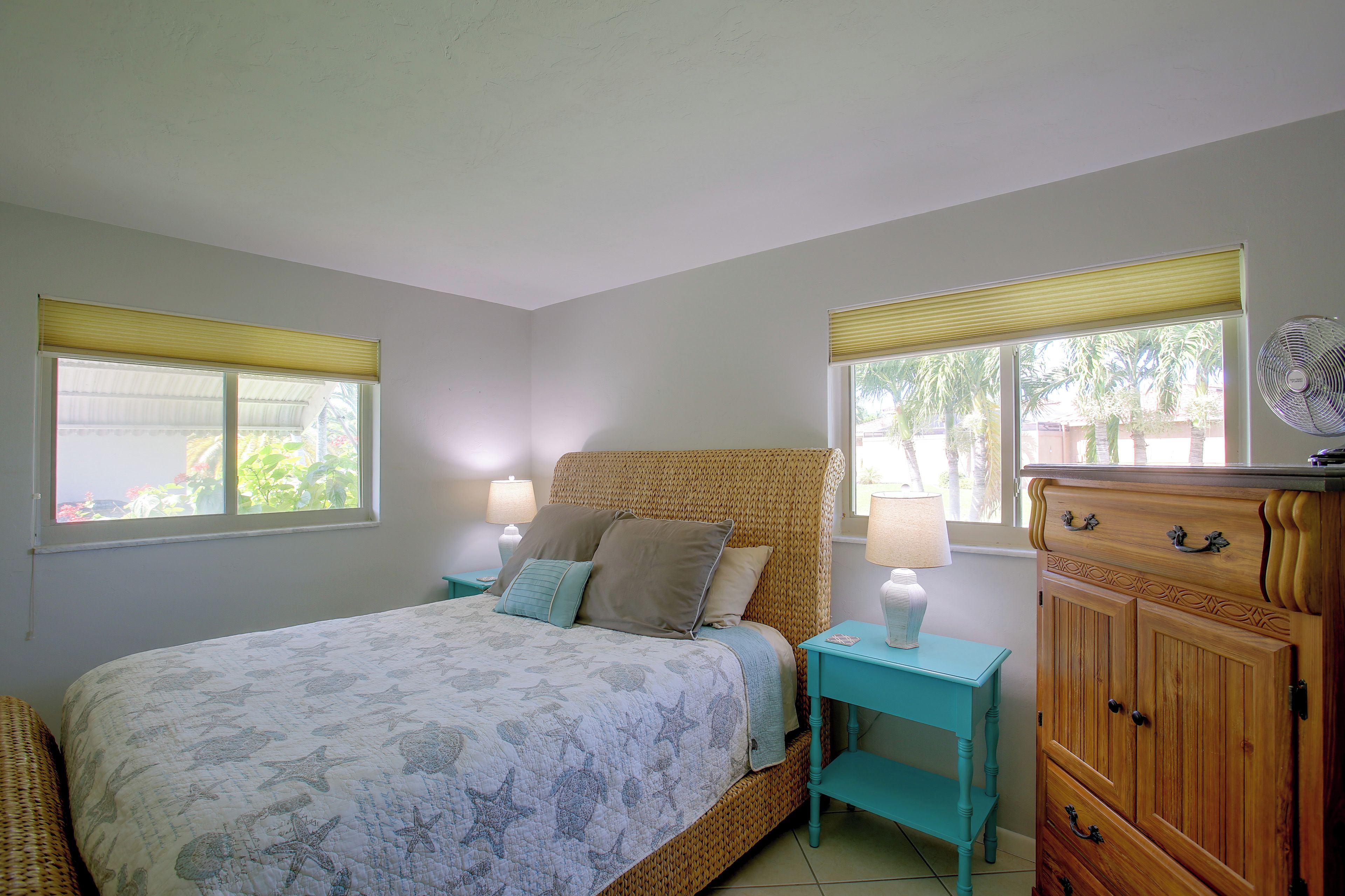 A bedroom with a bed, two bedside tables, a nightstand, a wooden cabinet, and windows with blinds.
