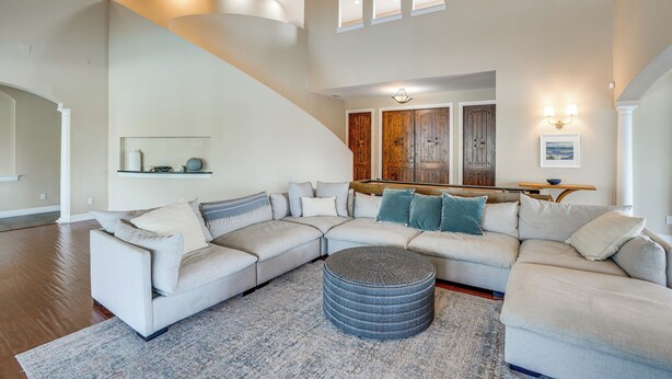 A spacious living room with a large L-shaped sectional sofa, a round ottoman, and a high ceiling with skylights.