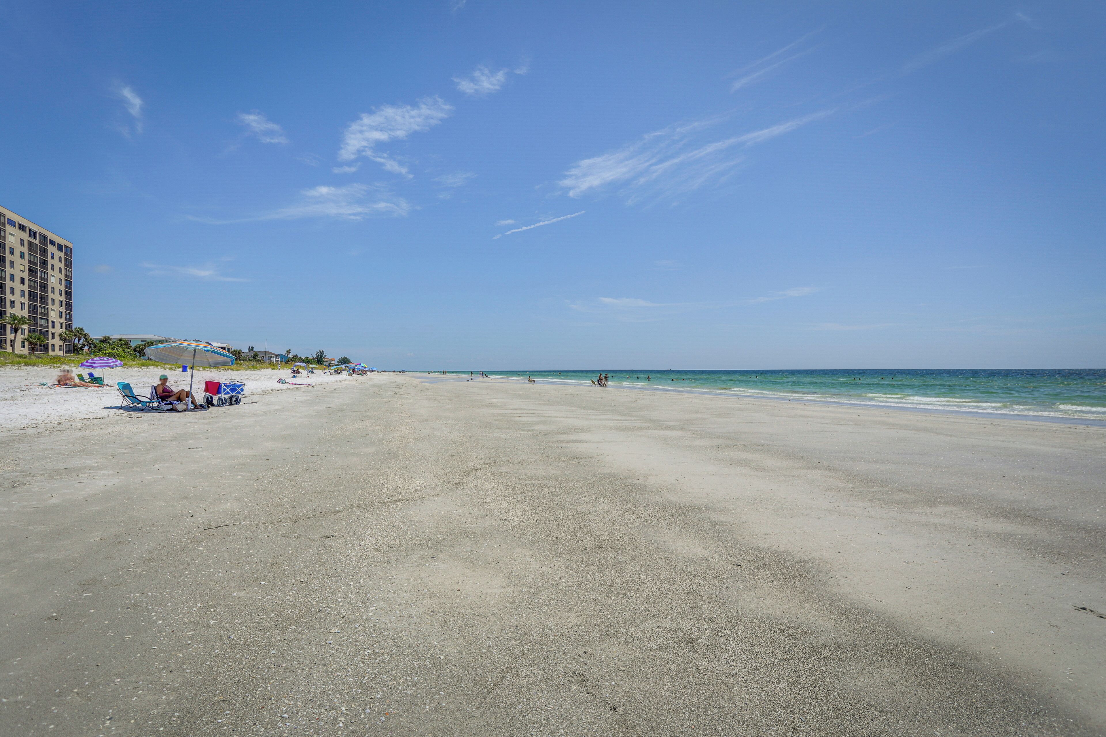 A wide sandy beach with a few people and beach chairs. There is a building on the left side.