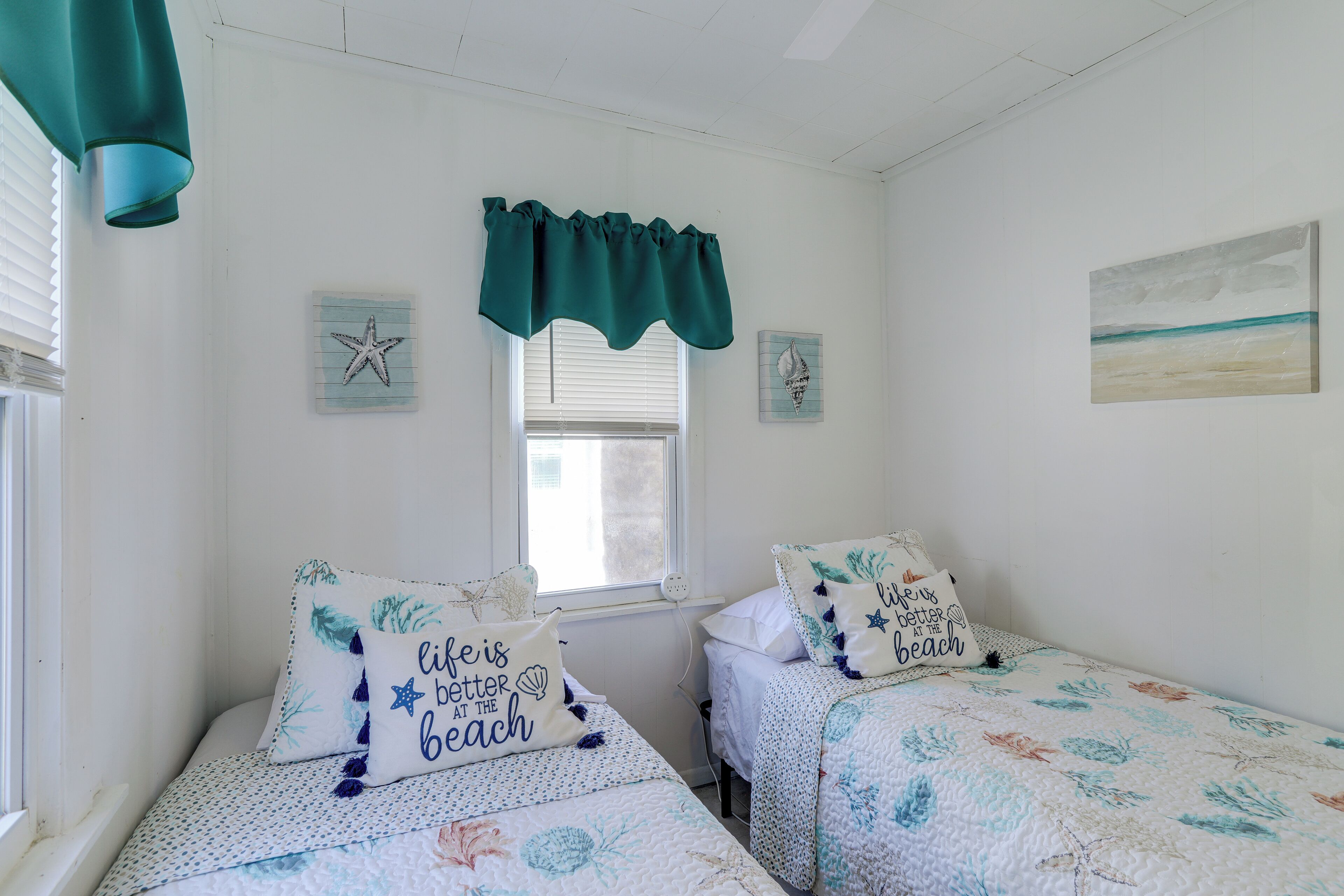 A bedroom with two beds, each with a floral bedspread and a pillow with a beach-themed design. There is a window with a white valance, and a painting of a beach scene on the wall.