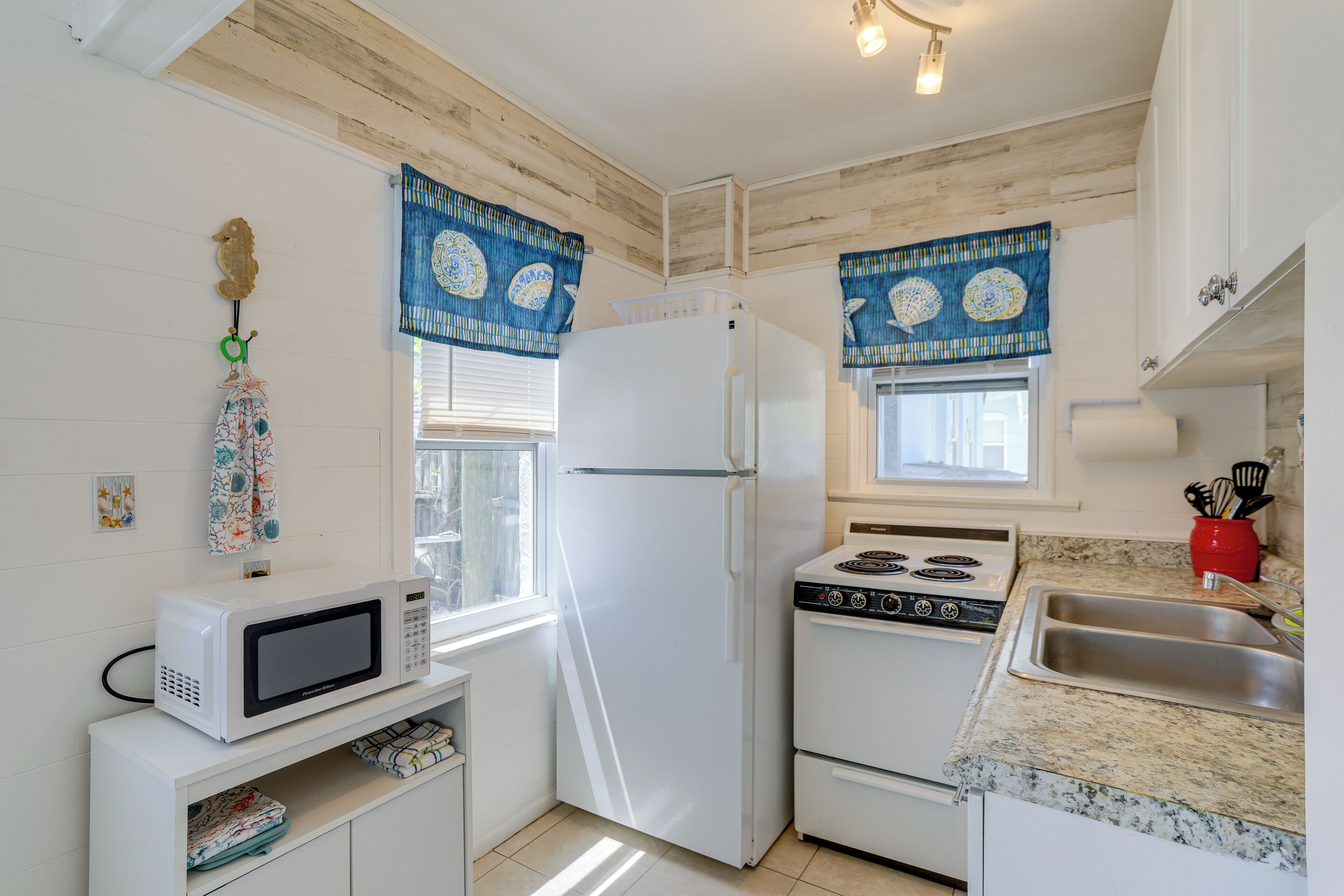 Cocina compacta con electrodomésticos blancos, encimera de granito y adornos de ventana azules con estampado.