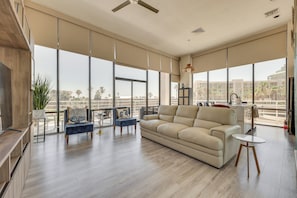 A spacious living room with a large beige sofa, a blue armchair, and a small round table. There is a large window providing a view of palm trees and buildings. - South Padre Island Gem: Community Pool & Hot Tub! (South Padre Island)