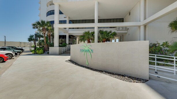 A modern building with a prominent logo, a parking area, and palm trees. - South Padre Island Gem: Community Pool & Hot Tub! (South Padre Island)