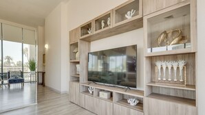 A modern living room with a large flat-screen TV, wooden shelving with decorative items, and a glass door leading to an outdoor seating area. - South Padre Island Gem: Community Pool & Hot Tub! (South Padre Island)