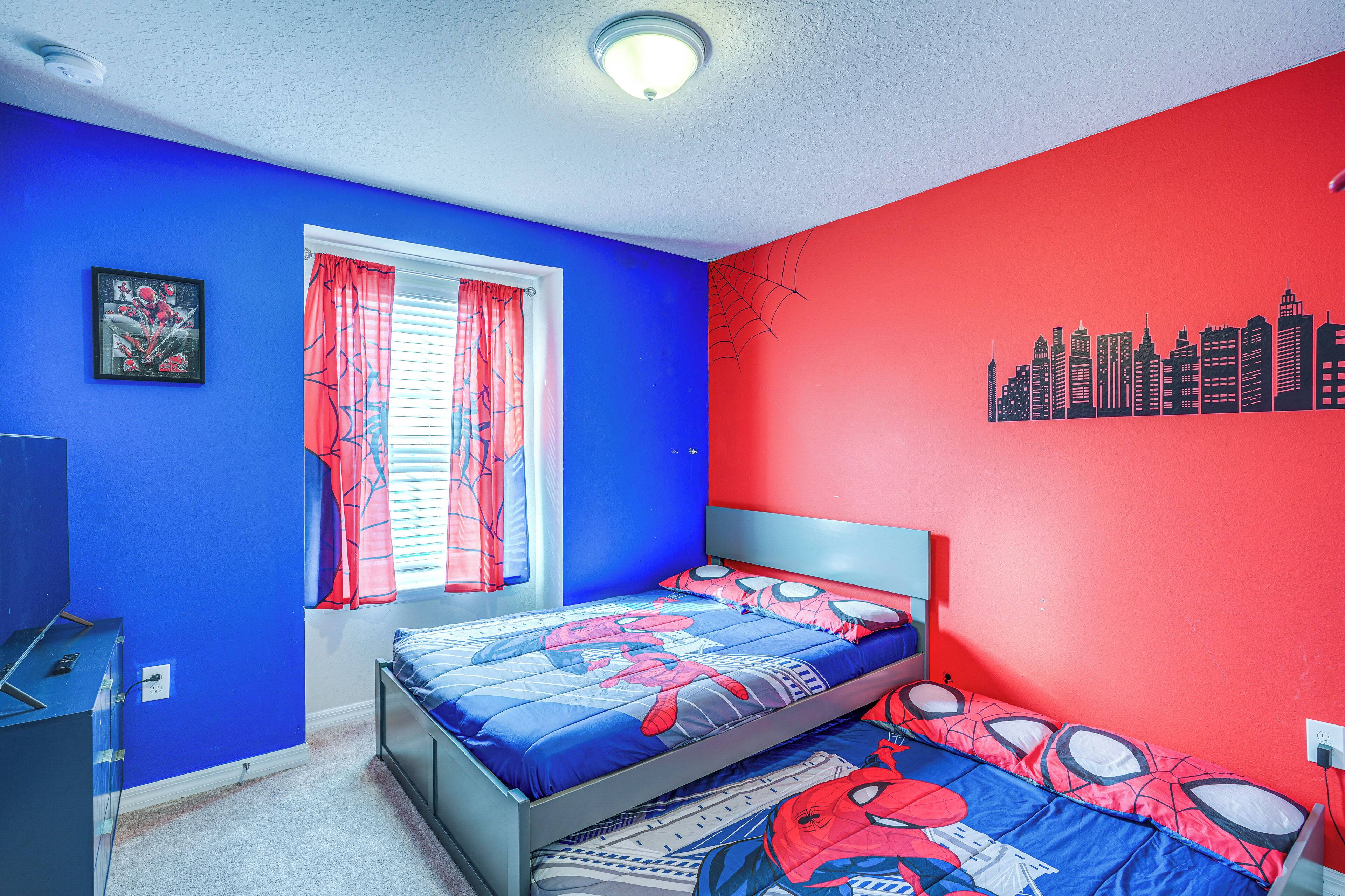 A bedroom with two beds, a window with red curtains, and a cityscape wall mural.