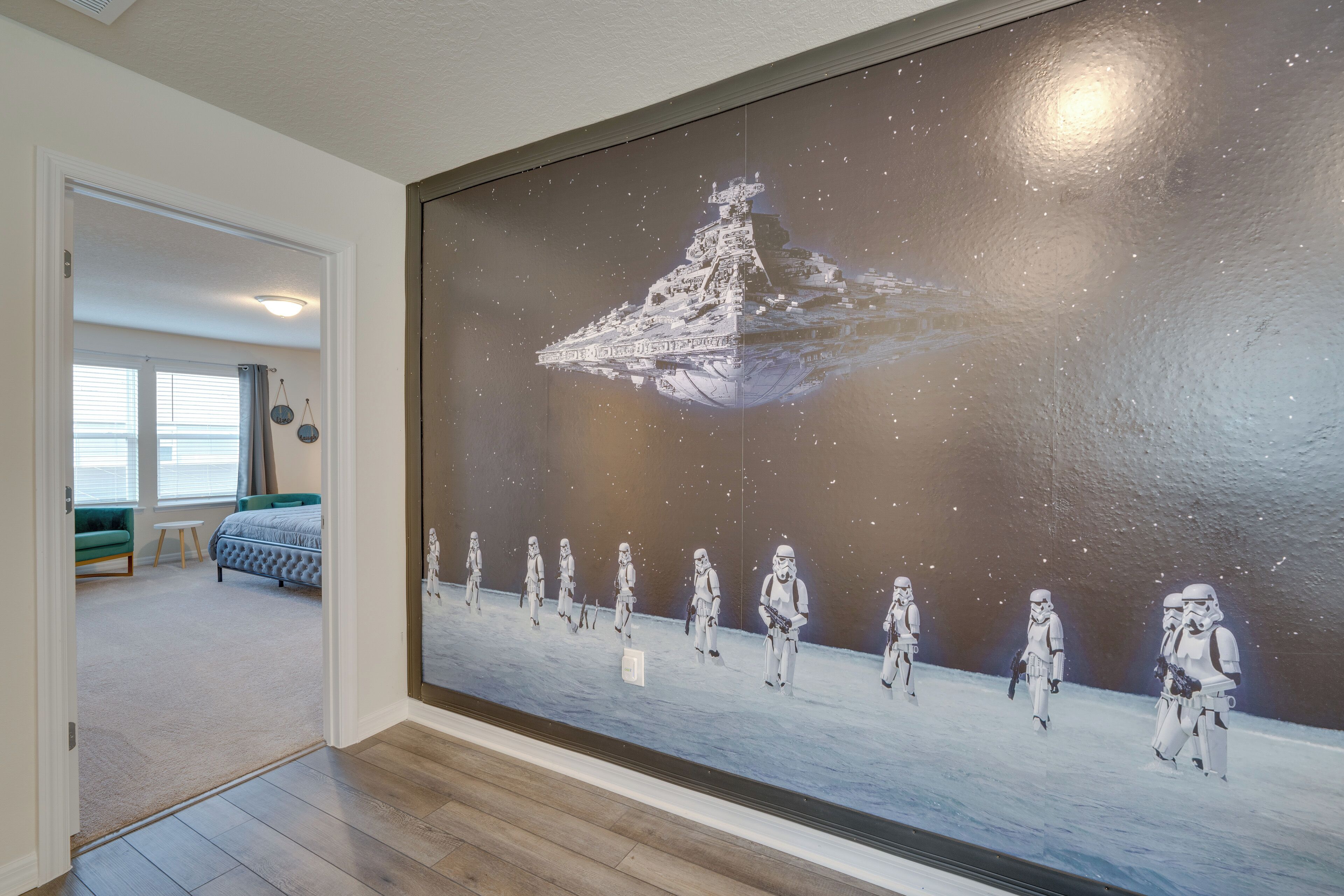 A bedroom with a large wall mural of a spaceship and stormtroopers, a bed, a green chair, and a window with curtains.