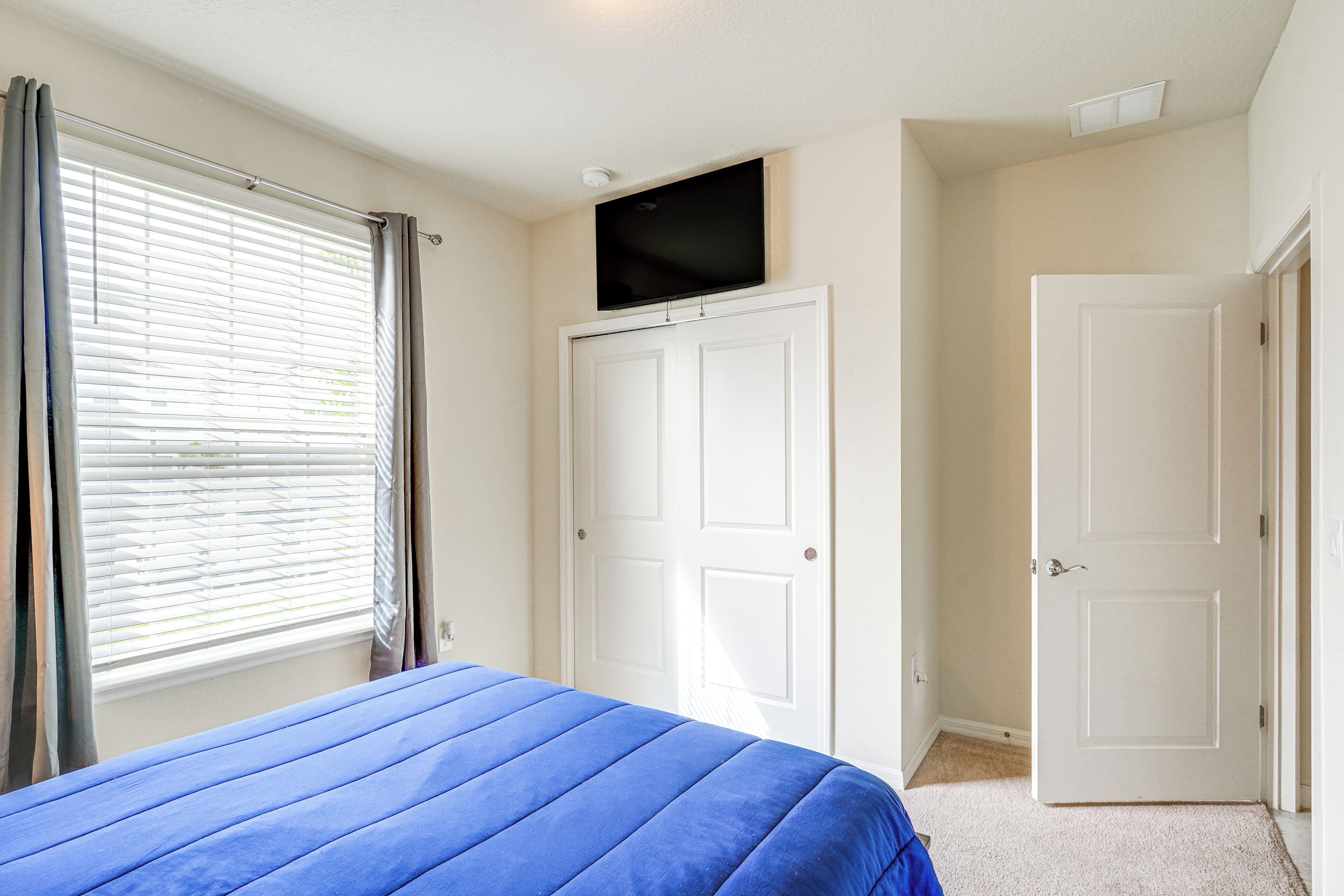A bedroom with a bed, a window with curtains, a white door, and a mounted television.