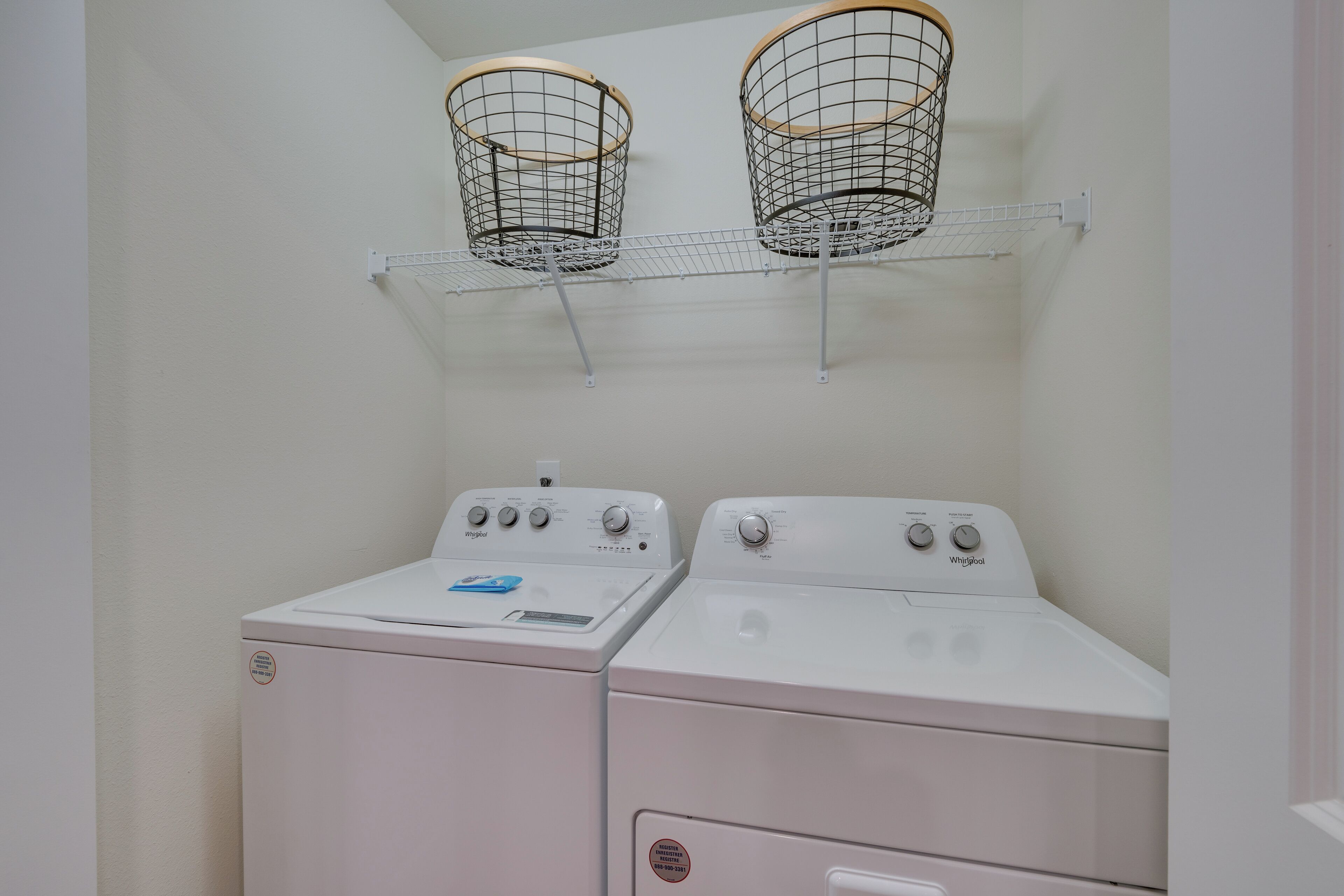 A laundry room with two white front-loading washers and dryers, a metal shelf with two baskets, and a white wall.