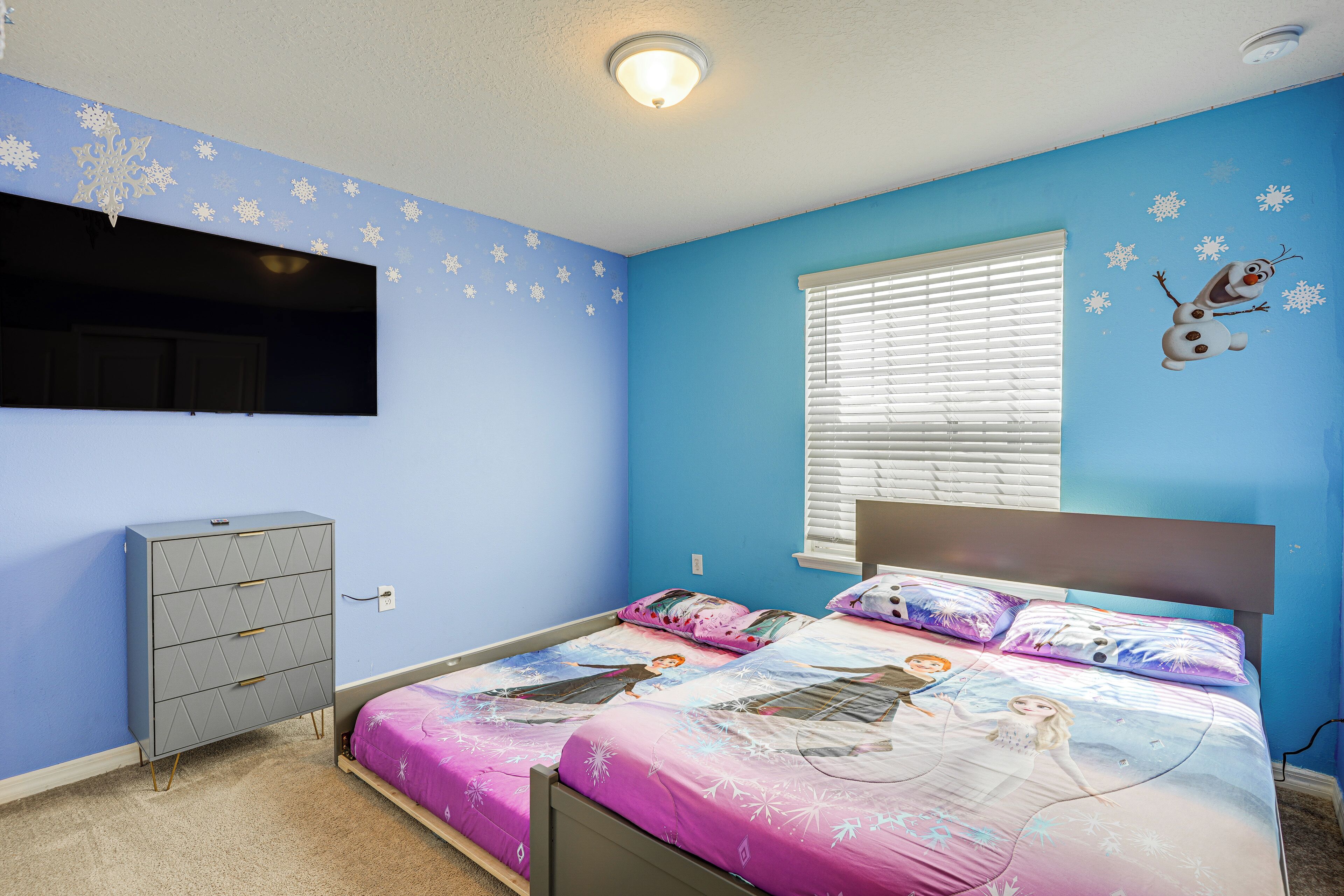 A bedroom with a bed featuring a Frozen-themed bedspread, a dresser, a window with blinds, and snowflake wall decals.