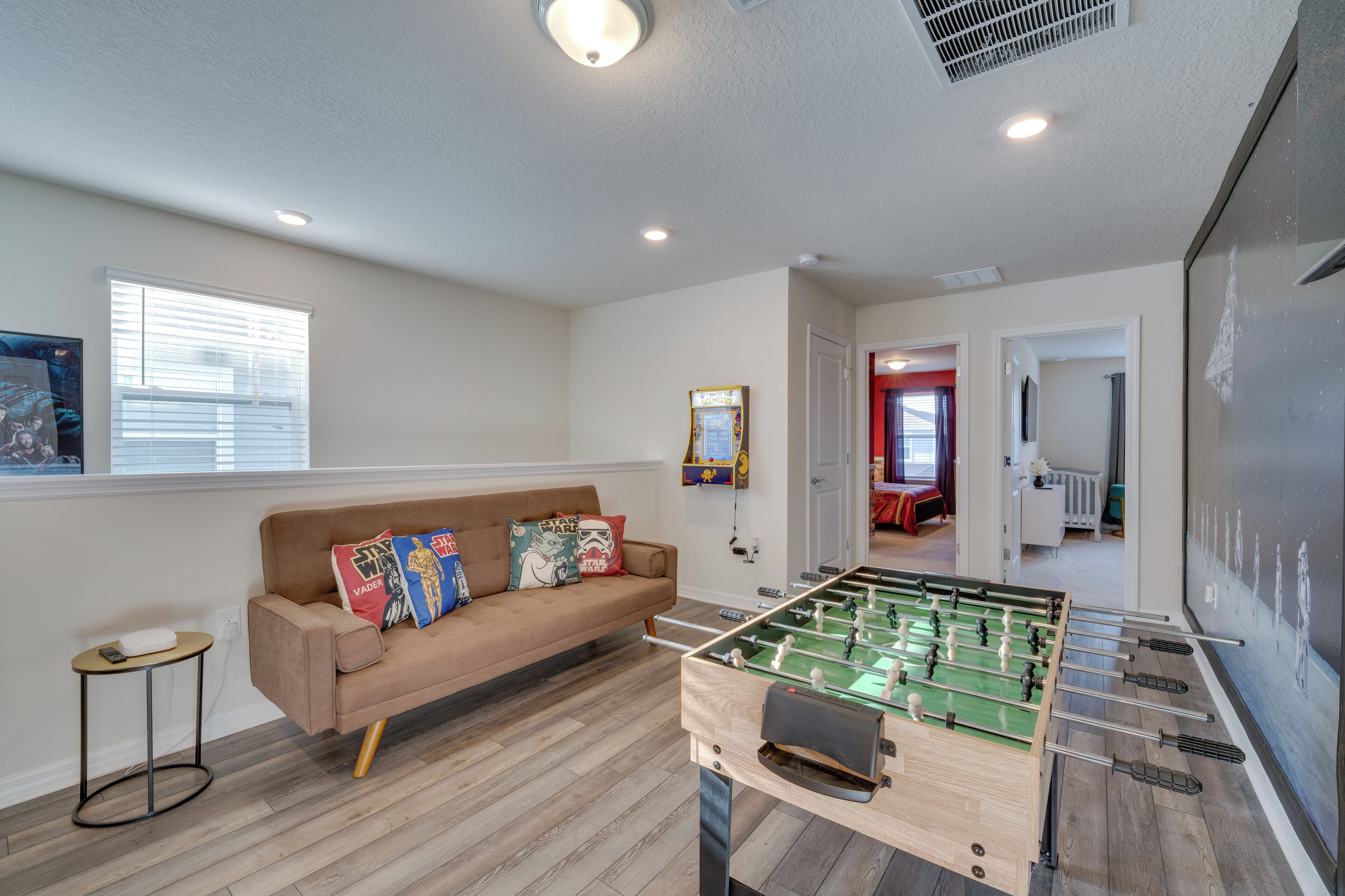 A room with a foosball table, a sofa with colorful pillows, a small side table, and a window with blinds.