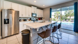 A modern kitchen with a stainless steel refrigerator, microwave, and coffee maker, a large island with bar stools, and a view of a swimming pool through large sliding glass doors. - Spacious Kissimmee Escape, Close to Disney World! (Kissimmee)