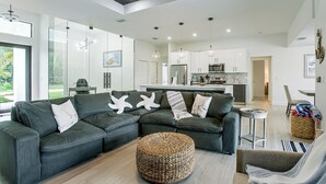 A modern living room with a sectional sofa, a woven ottoman, and a dining area with a kitchen in the background. - Private Pool & Grill: Cape Coral Vacation Rental! (Cape Coral)