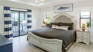 A bedroom with a large bed, a ceiling fan, a view to the outside, and a dining area with a grill. - Private Pool & Grill: Cape Coral Vacation Rental! (Cape Coral)