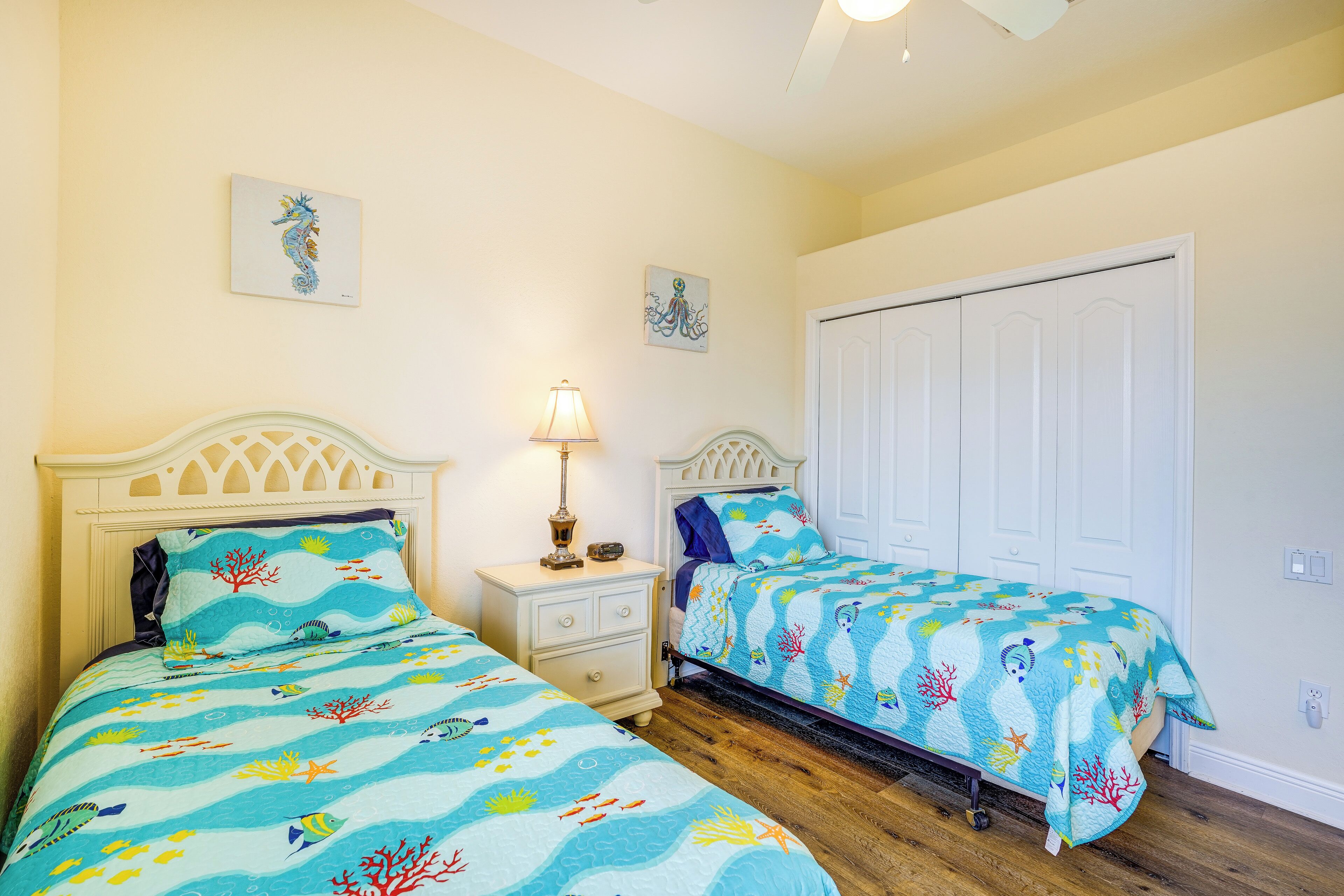 A bedroom with two beds, each with a blue ocean-themed bedspread. There is a nightstand with a lamp between the beds, and a white wardrobe against the wall.