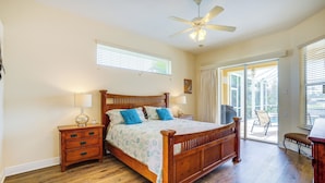A bedroom with a wooden bed, a nightstand, a lamp, a ceiling fan, and a view of the outdoors through a sliding glass door.