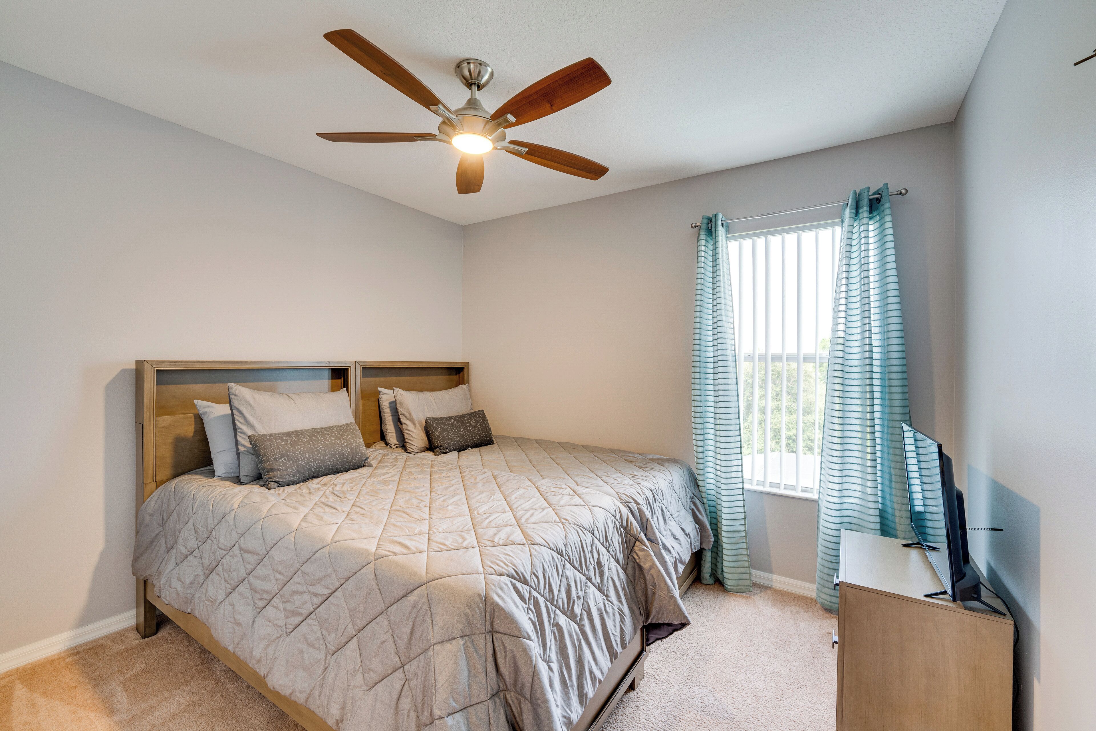 A bedroom with a bed, a ceiling fan, a window with curtains, and a desk with a computer.