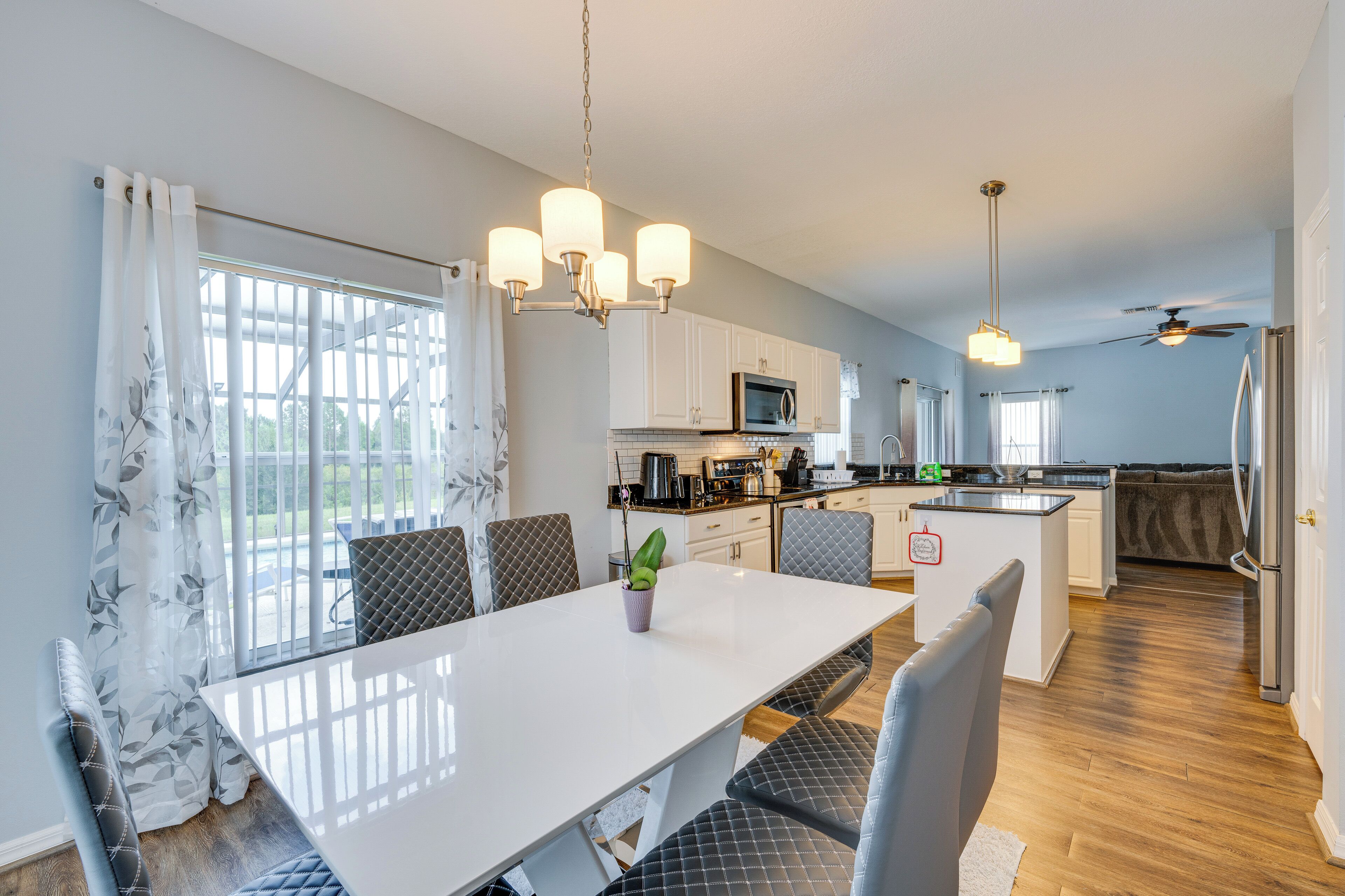A modern kitchen with white cabinets, a central island, and a dining area with a white table and grey upholstered chairs.